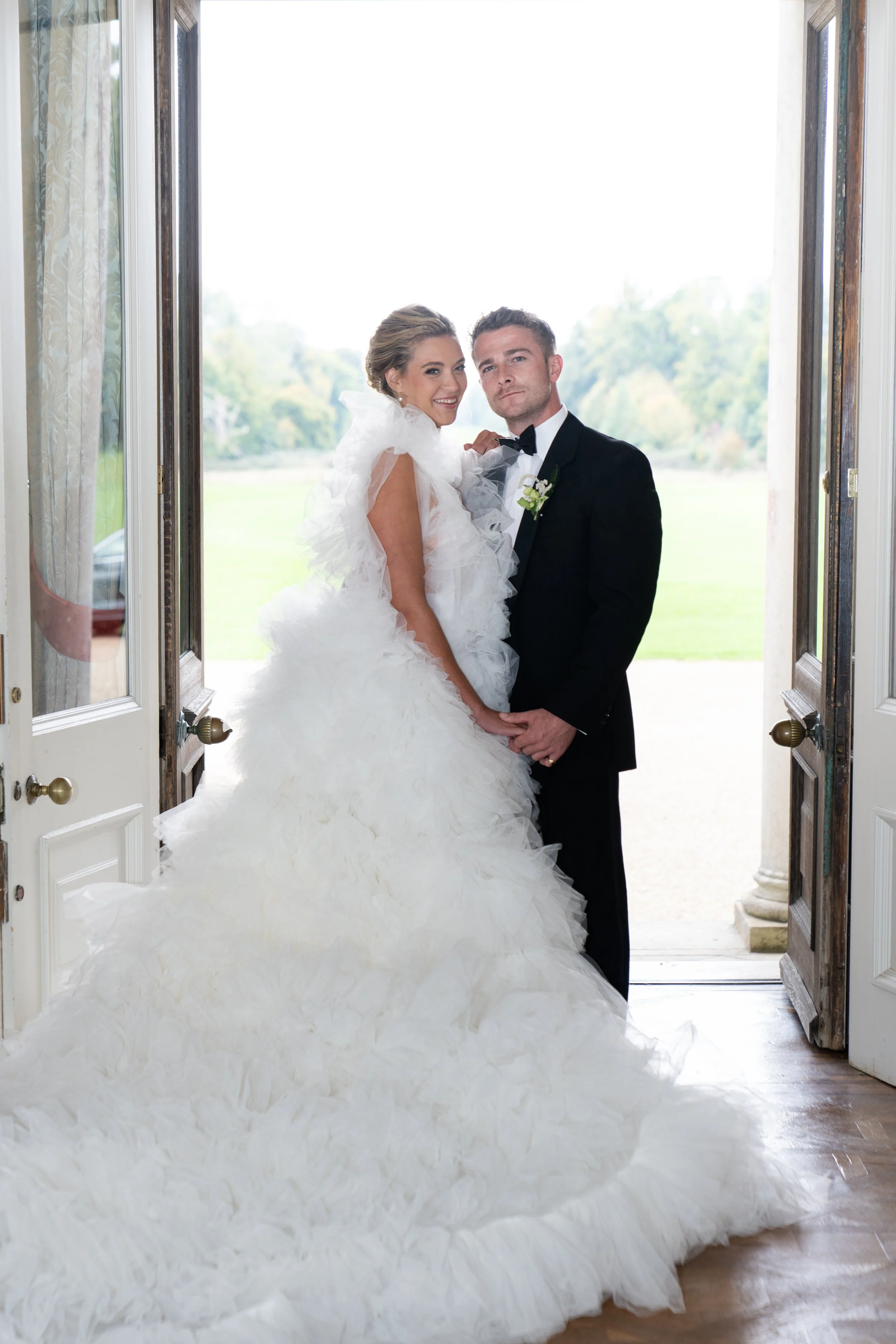 A bride in a white wedding gown and a groom in a black tuxedo standing together in a doorway with a view of a green outdoor landscape in the background.