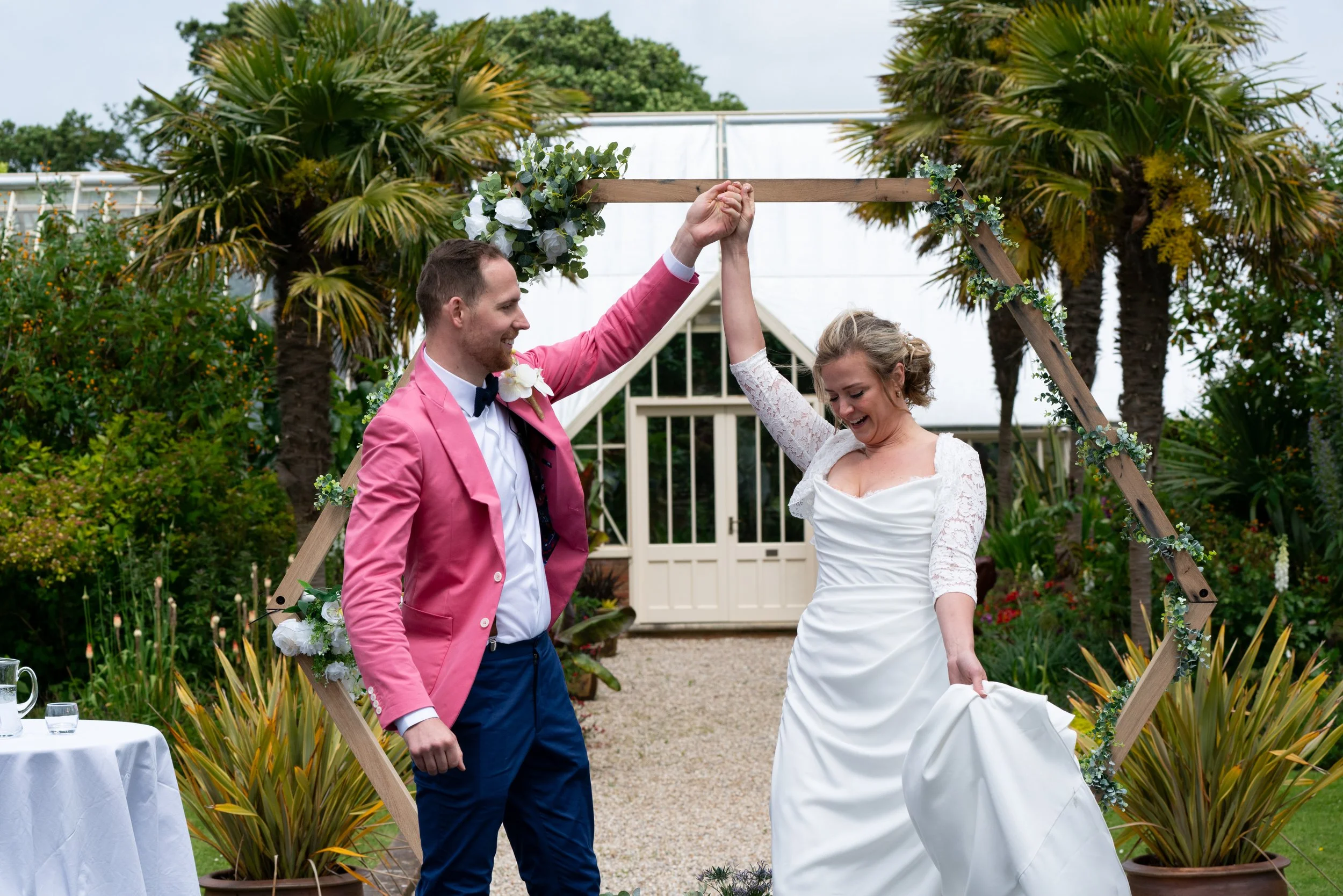 A couple celebrating their wedding ceremony, holding hands aloft and smiling and laughing, with tropical plants and a decorative arch in the background.