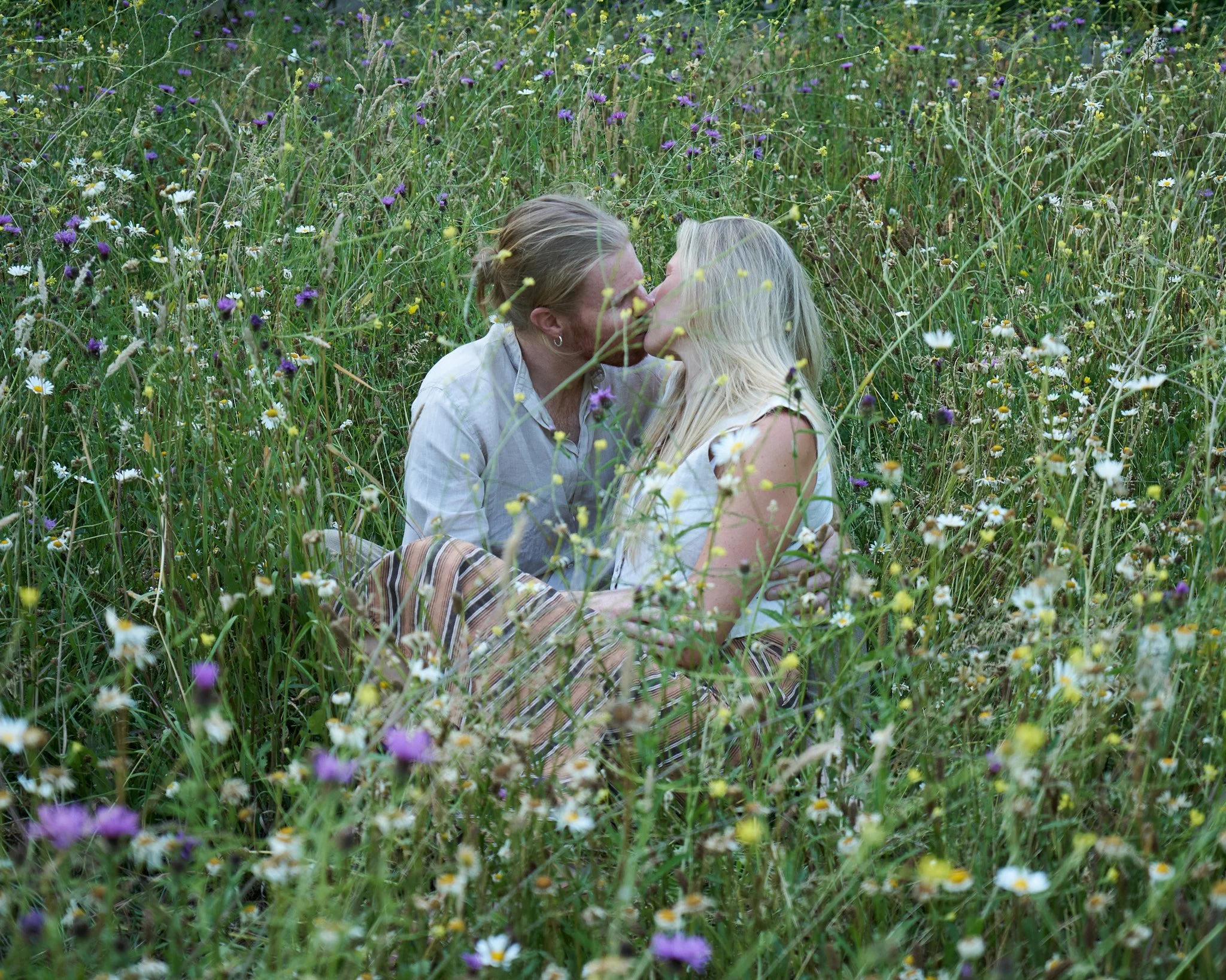 A couple sitting in a meadow of wildflowers sharing a kiss.