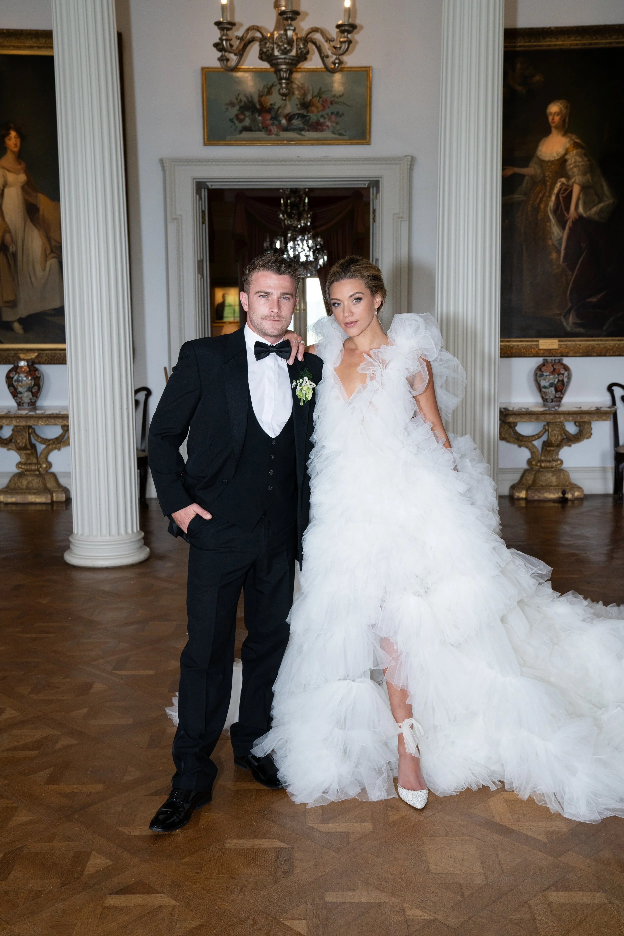 A bride and groom stand together in an elegant room with wooden floors, white columns, and large paintings. The groom wears a black tuxedo with a bow tie, and the bride is in a white, ruffled wedding gown with tulle sleeves.