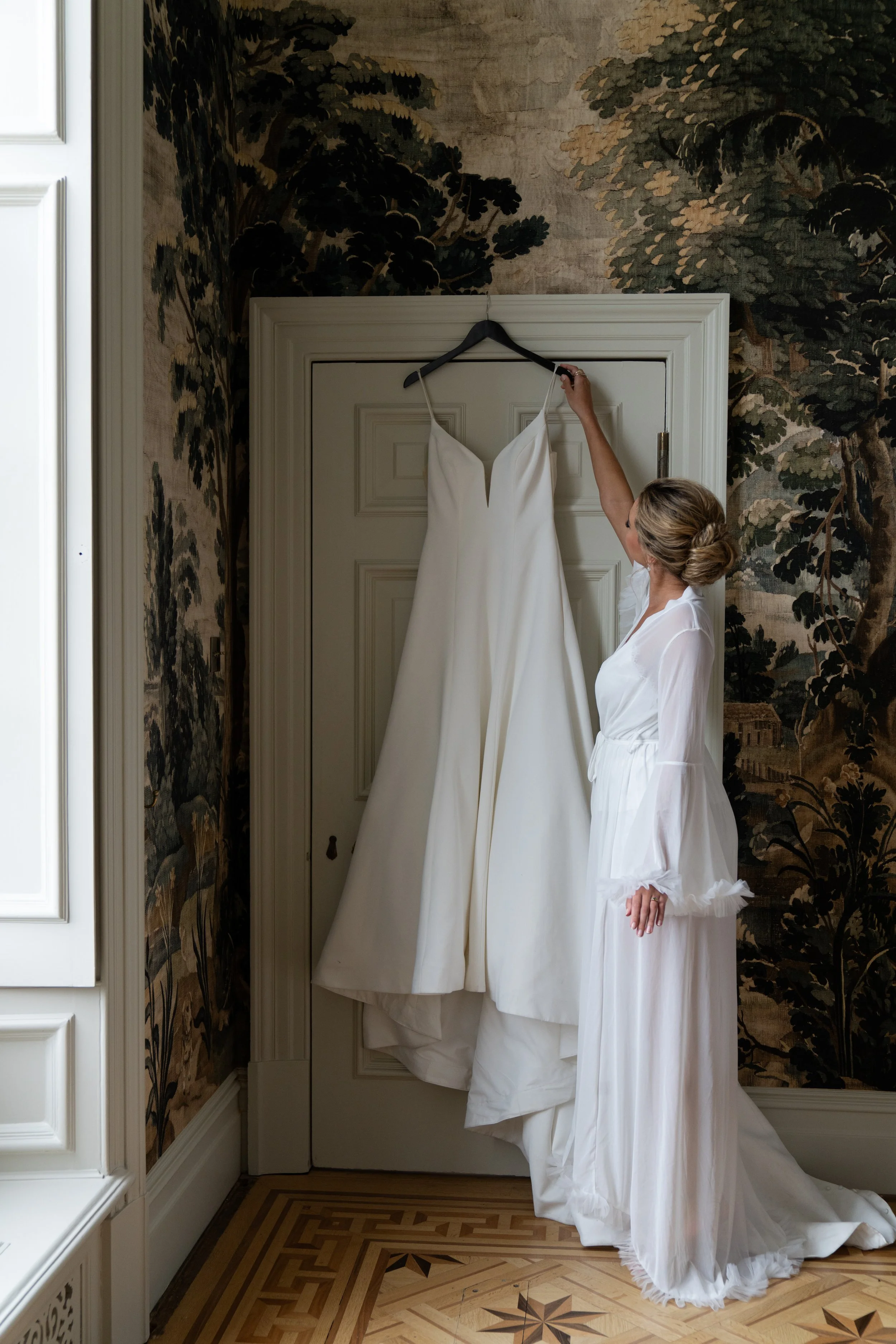 A woman in a white robe looking at a white wedding dress hanging on a hanger in a room with ornate wallpaper and a patterned wood floor.