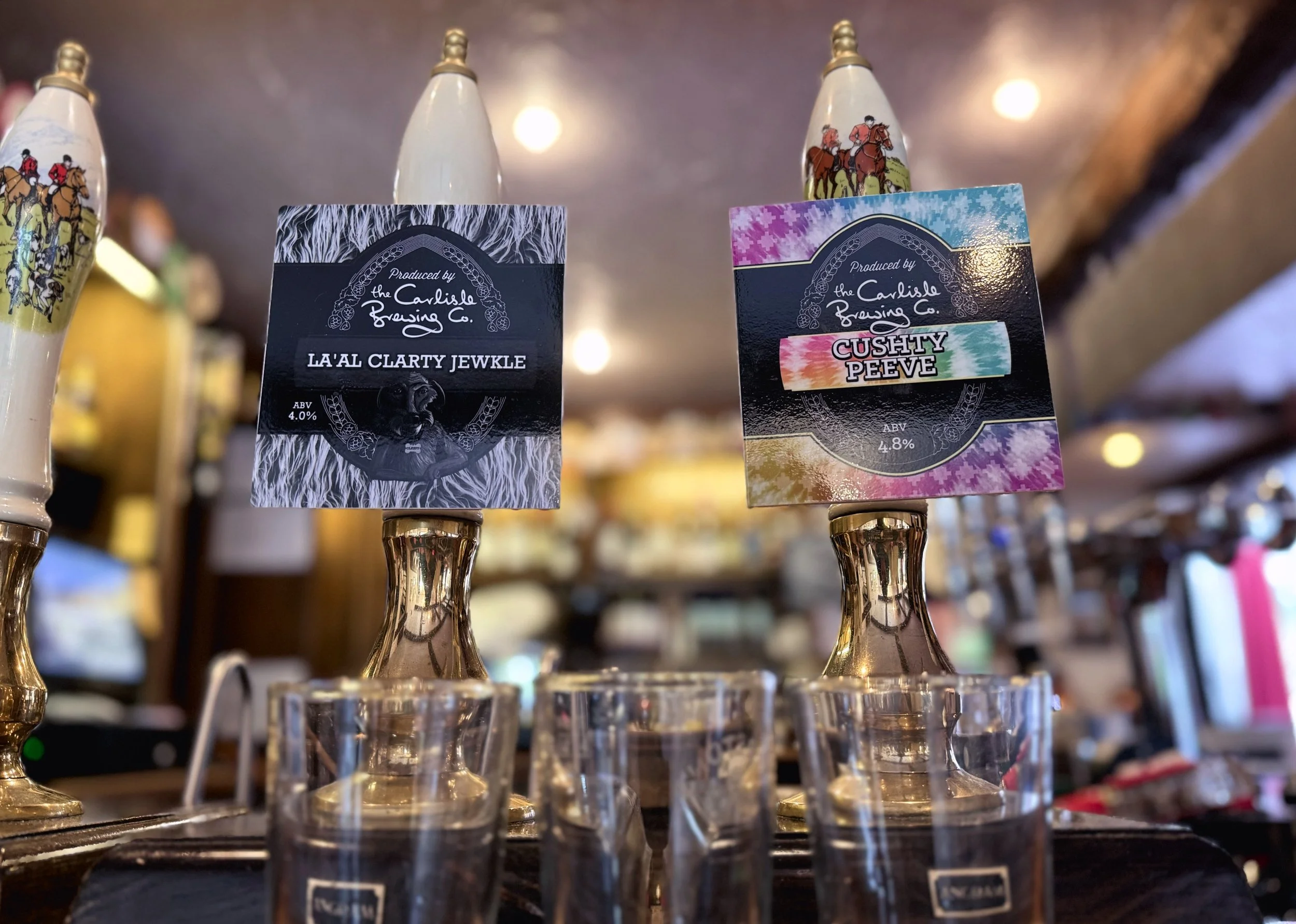 Two beer tap handles with colorful labels in a bar. The left handle has a black-and-white label with a lion illustration, reading "LA'AL CLARTY JEWKLE" with 4.0% ABV. The right handle has a vibrant, tie-dye style label that says "CUSHY PEEVE" with 4.8% ABV. Glasses are placed beneath the taps.