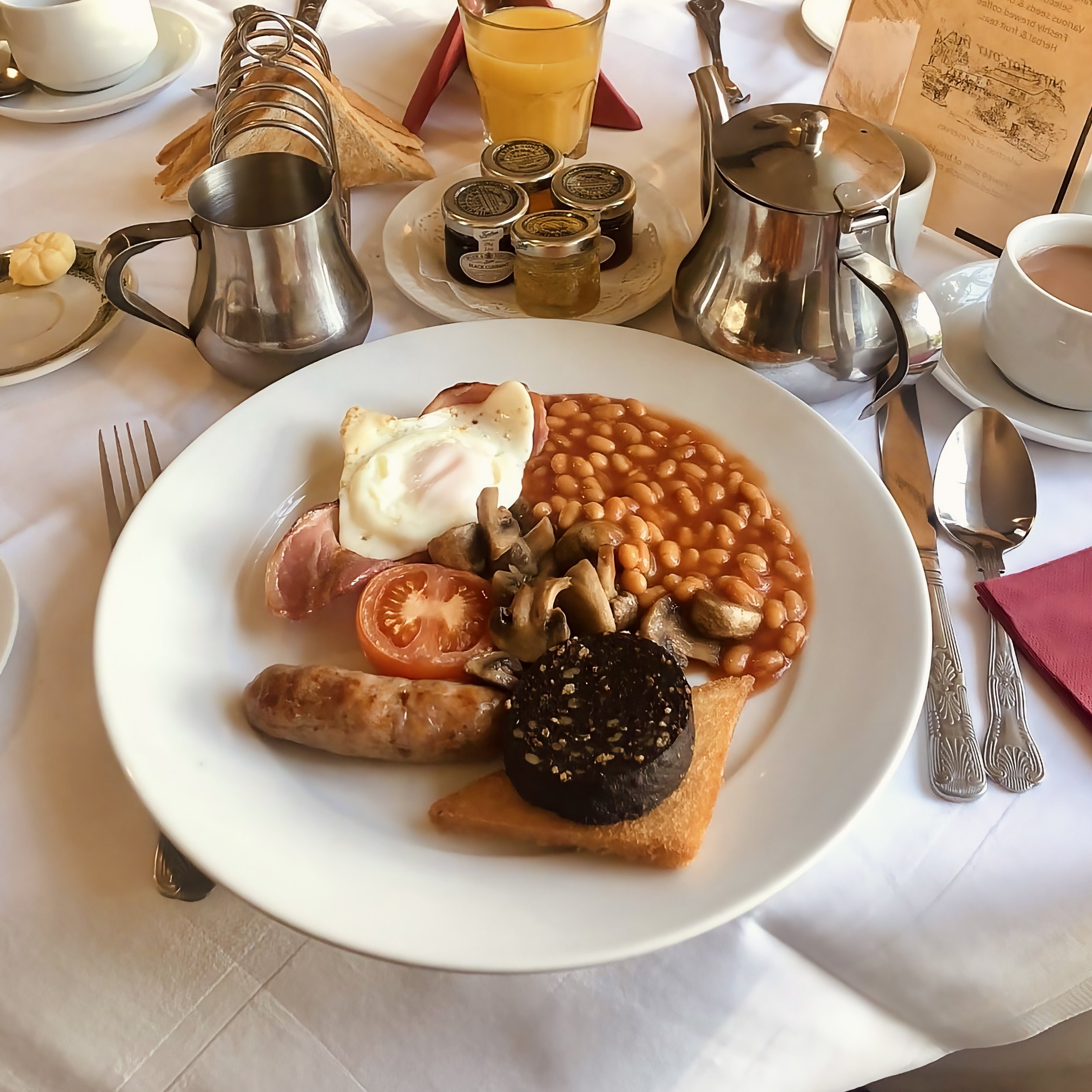 A breakfast plate with baked beans, mushrooms, tomato, poached egg, sausage, black pudding, and toast on a table set for breakfast.