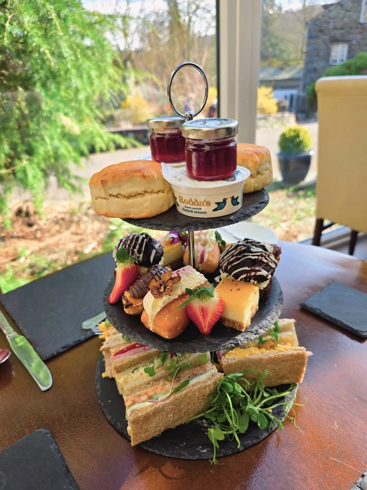 Three-tiered platter with scones, jam, cream, finger sandwiches, and assorted desserts, set on a wooden table near a window with a garden view.