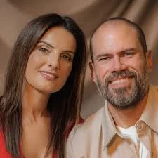 A smiling couple standing in front of a wooden door, with the man wearing a checked shirt and the woman in a white blouse with a black geometric pattern.