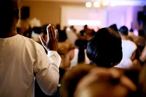People worshiping in a church with arms raised, facing a screen with blurred text, illuminated by colorful lights.