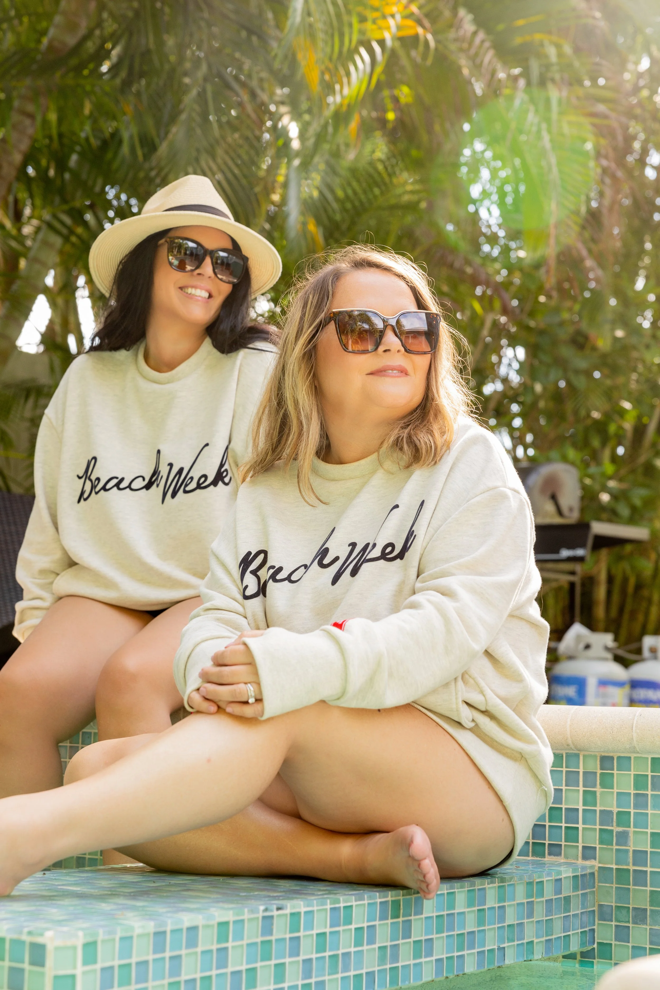 Two women in sunglasses and hats sitting by a pool, wearing "Beach Week" sweatshirts, with tropical plants in the background.