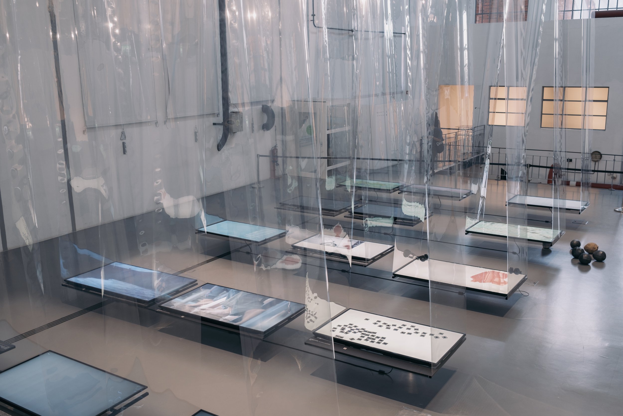 High perspective looking down at TV screens suspended from sheets of plastic in a gallery space.