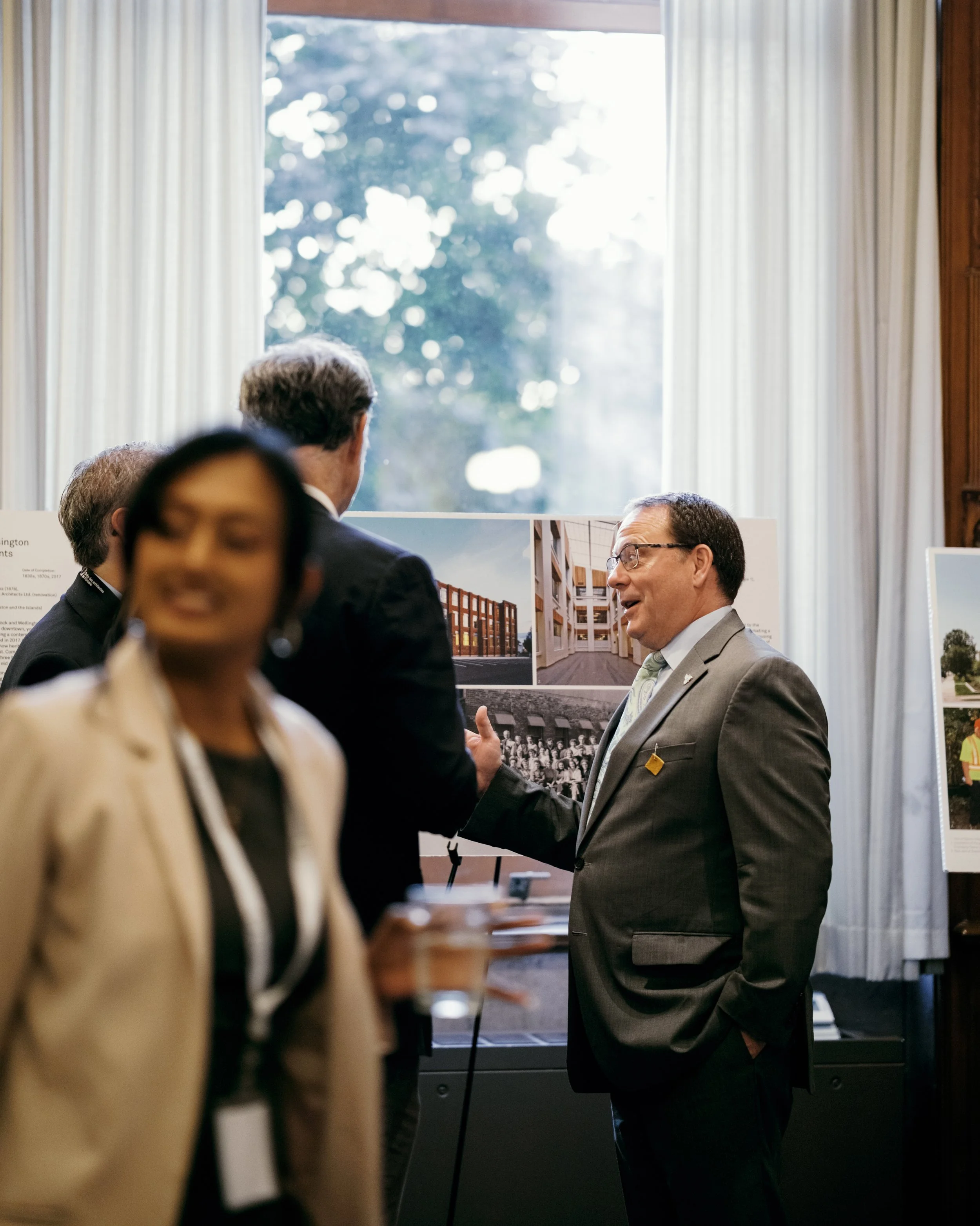 Mike Schreiner, MPP, speaking with architects at a reception in front of an exhibition board.
