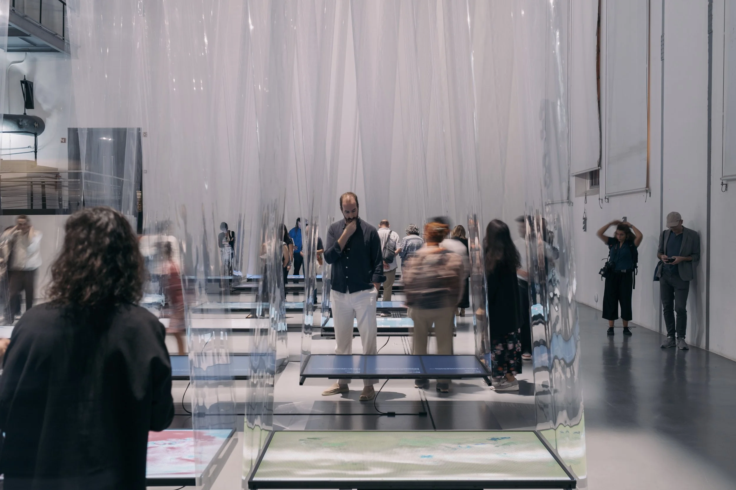 Two people view a film on a suspended horizontal screen in a gallery space. Other people are moving around the space around them.