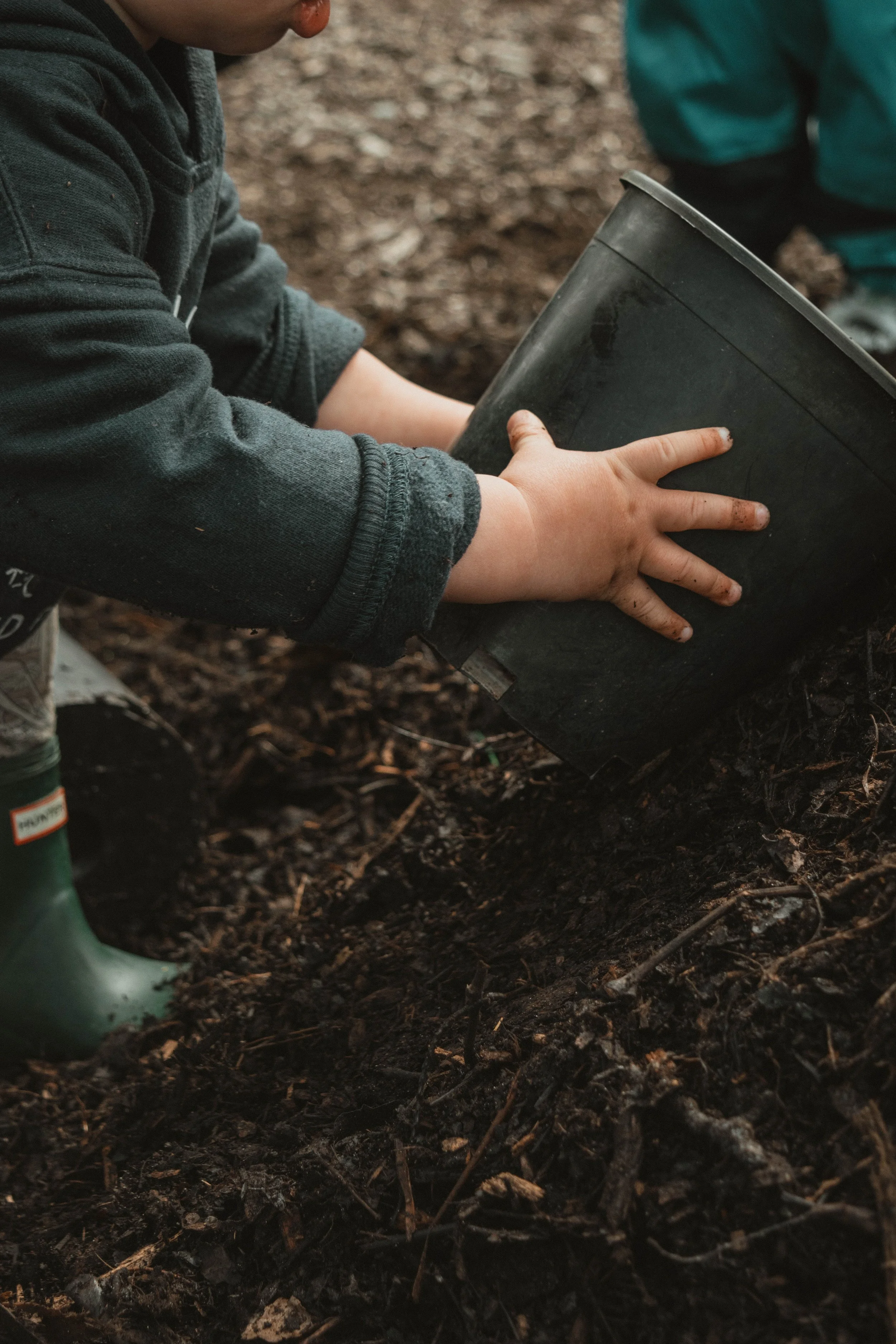 Child planting a seed in the soil using a black plastic container outdoors.