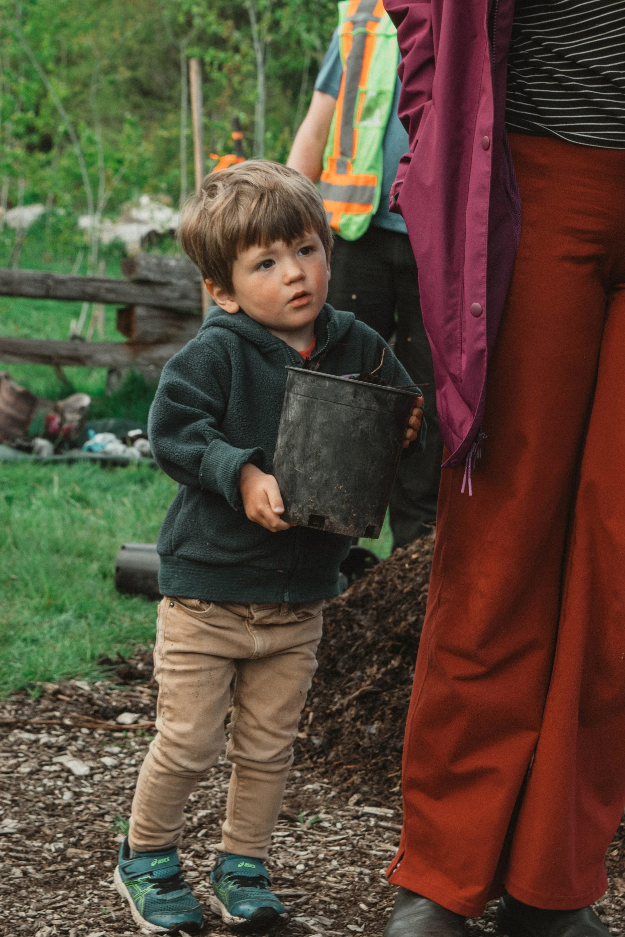 A young boy holding a small black bucket outside, surrounded by people in a lush, green outdoor setting.