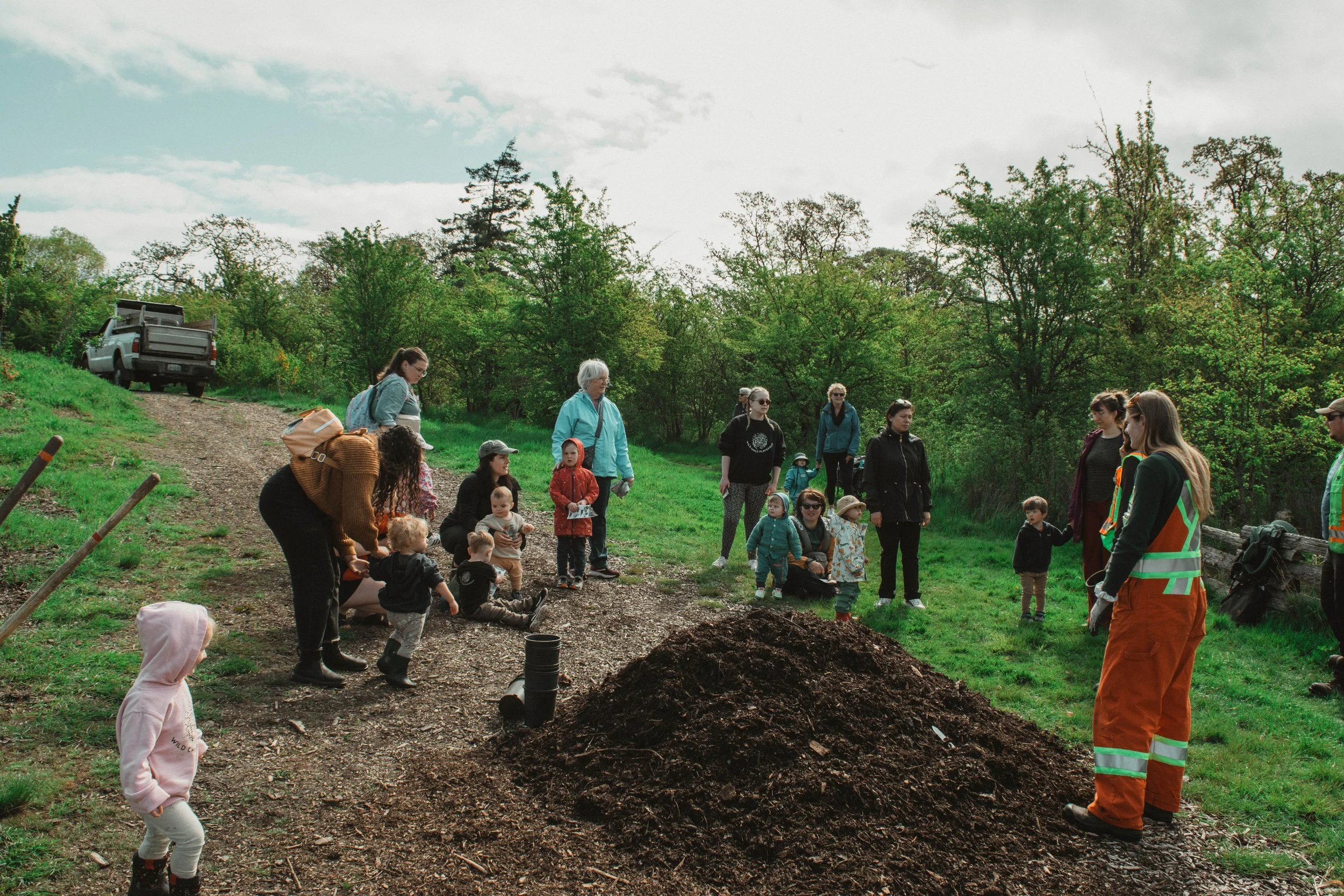 Group of children and adults gathered outdoors around a pile of soil, listening to a person in orange safety gear during a community or educational event.