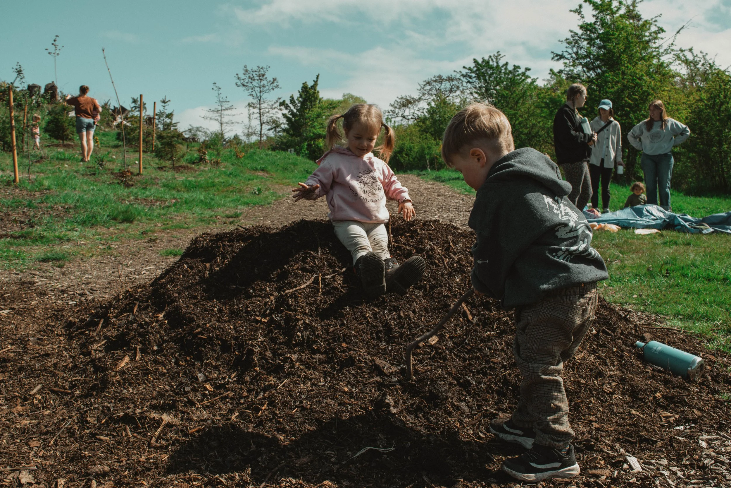 Children playing and digging in a mound of dirt outdoors on a sunny day with trees and a group of people in the background.