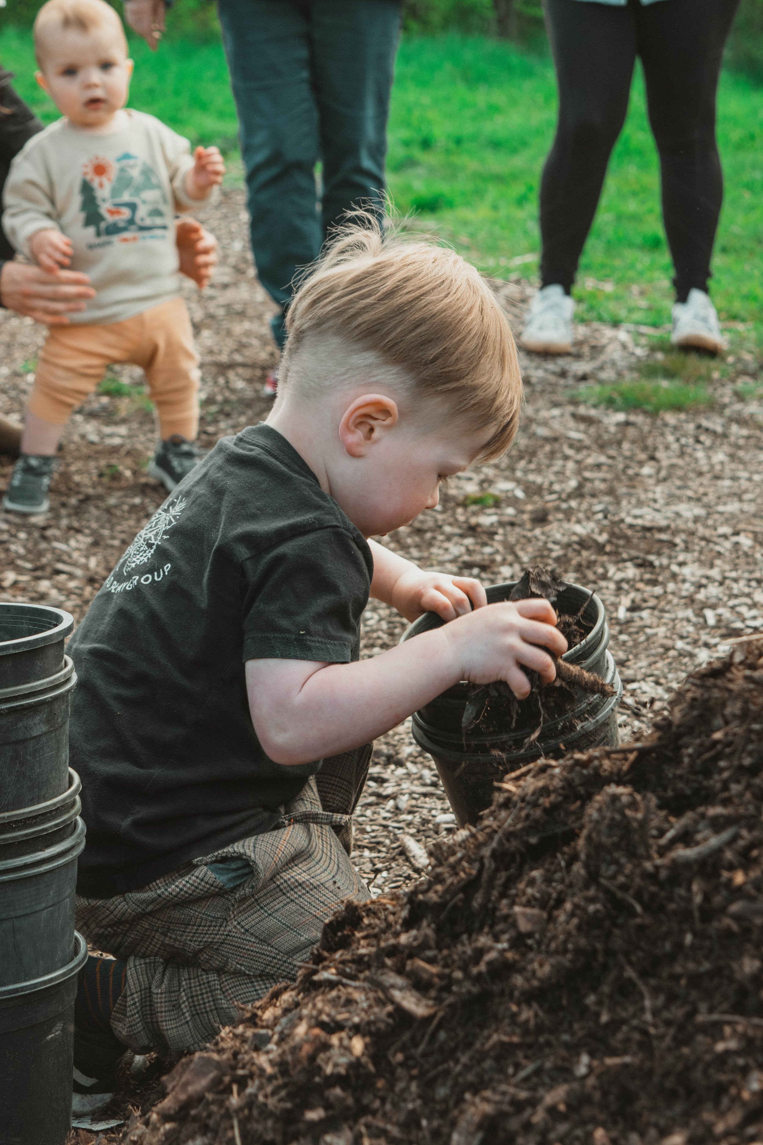A young boy planting a seed in a pot during an outdoor activity, with other children and adults nearby.
