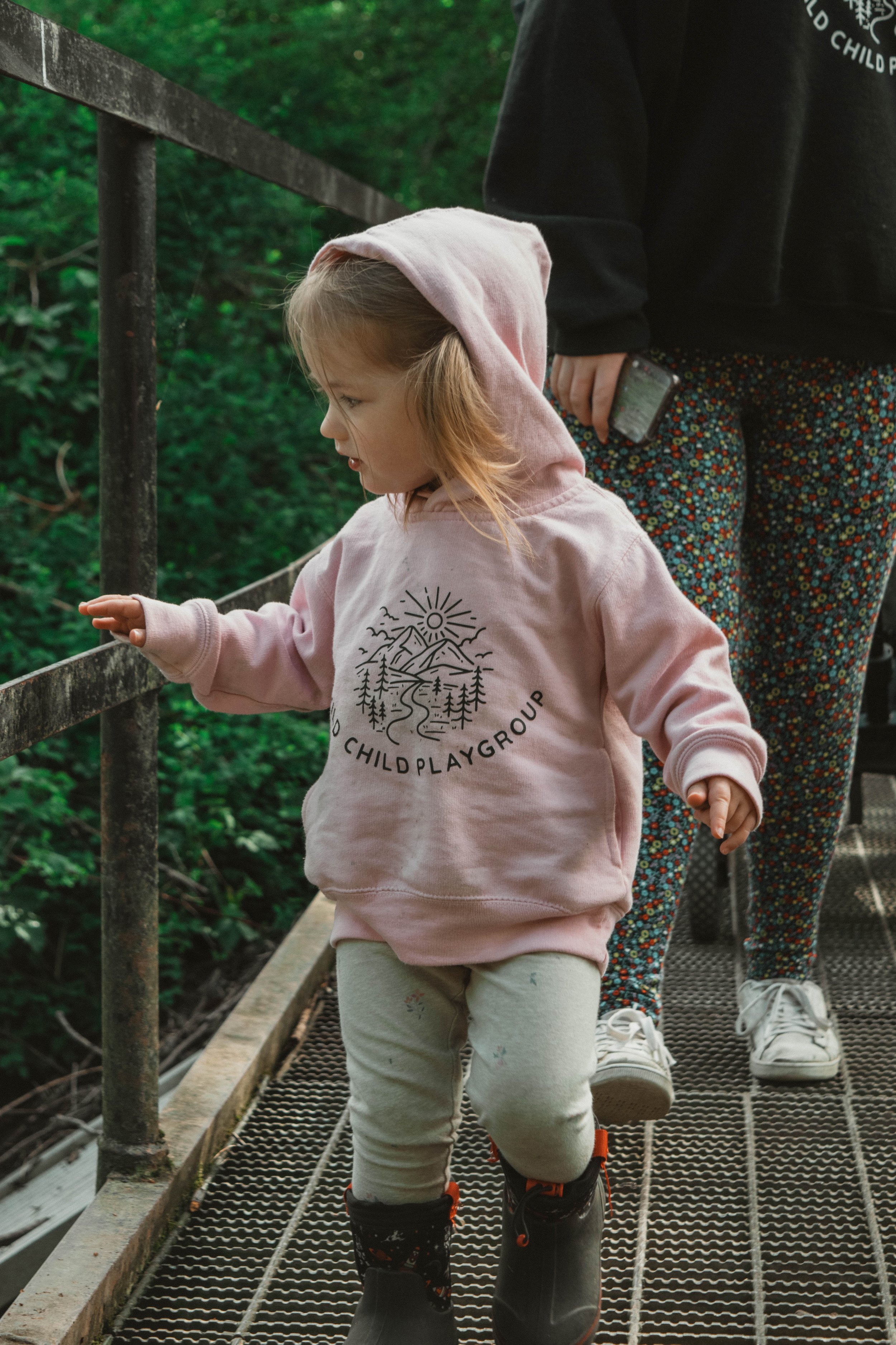 A young girl wearing a pink hoodie, grey pants, and black boots with orange laces, standing on a metal bridge, pointing at the railing while touring outdoors with an adult person in the background holding a smartphone, surrounded by green trees.