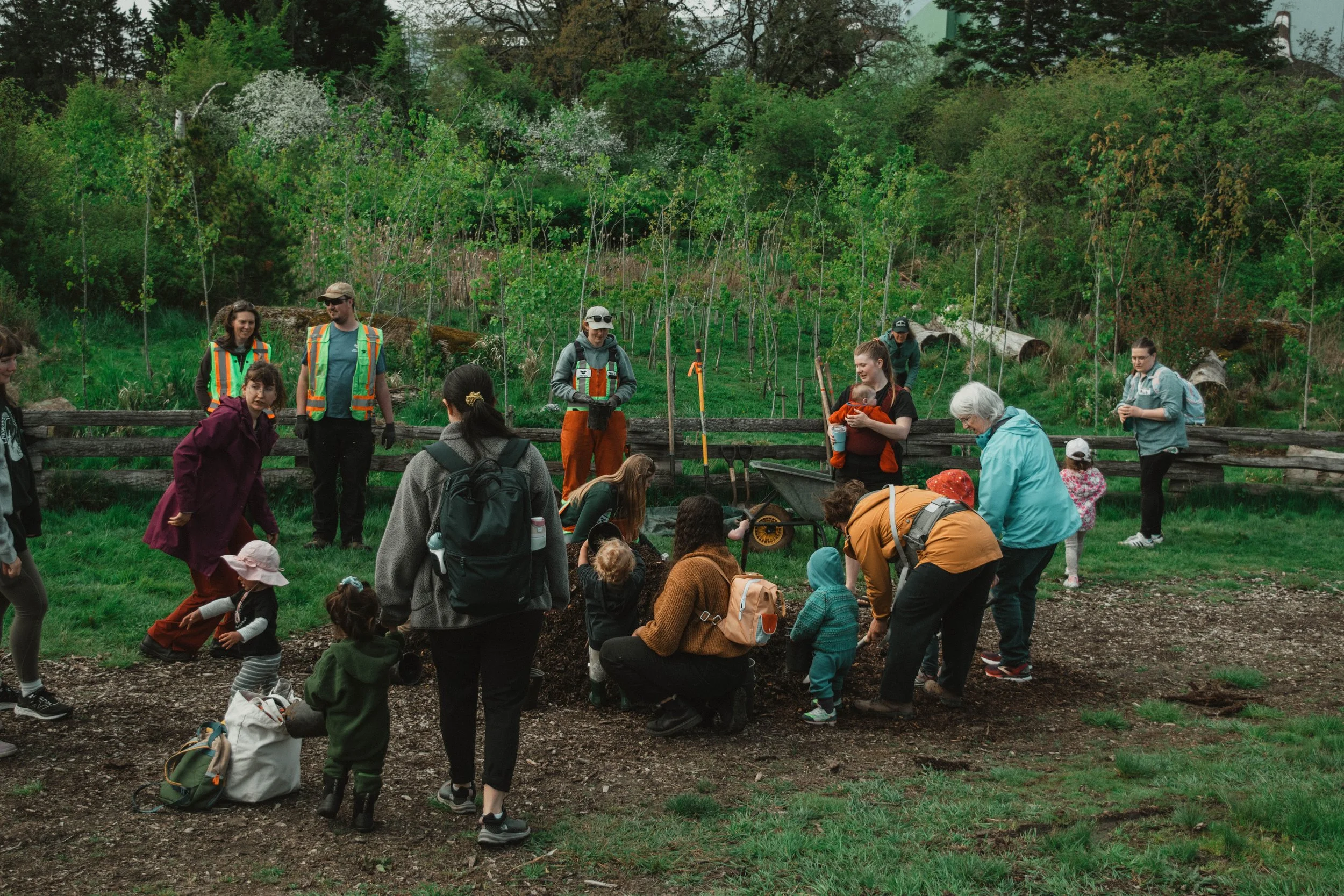 Group of people including children and adults gathered outdoors on a grassy area with trees in the background, some kneeling or bending down, engaged in an activity or discussion.