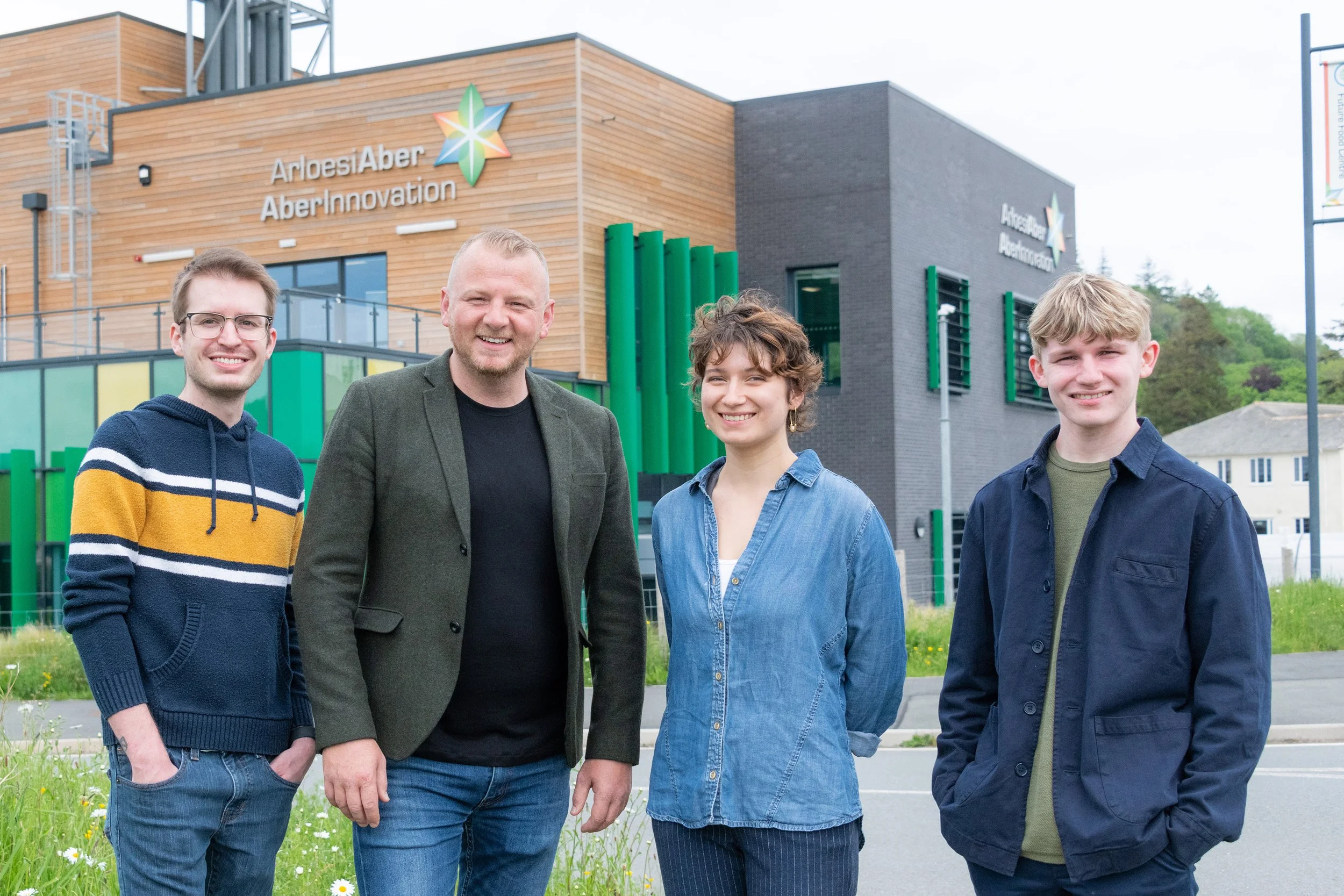 Four people standing outdoors in front of the Aber Aber Innovation building, smiling at the camera.