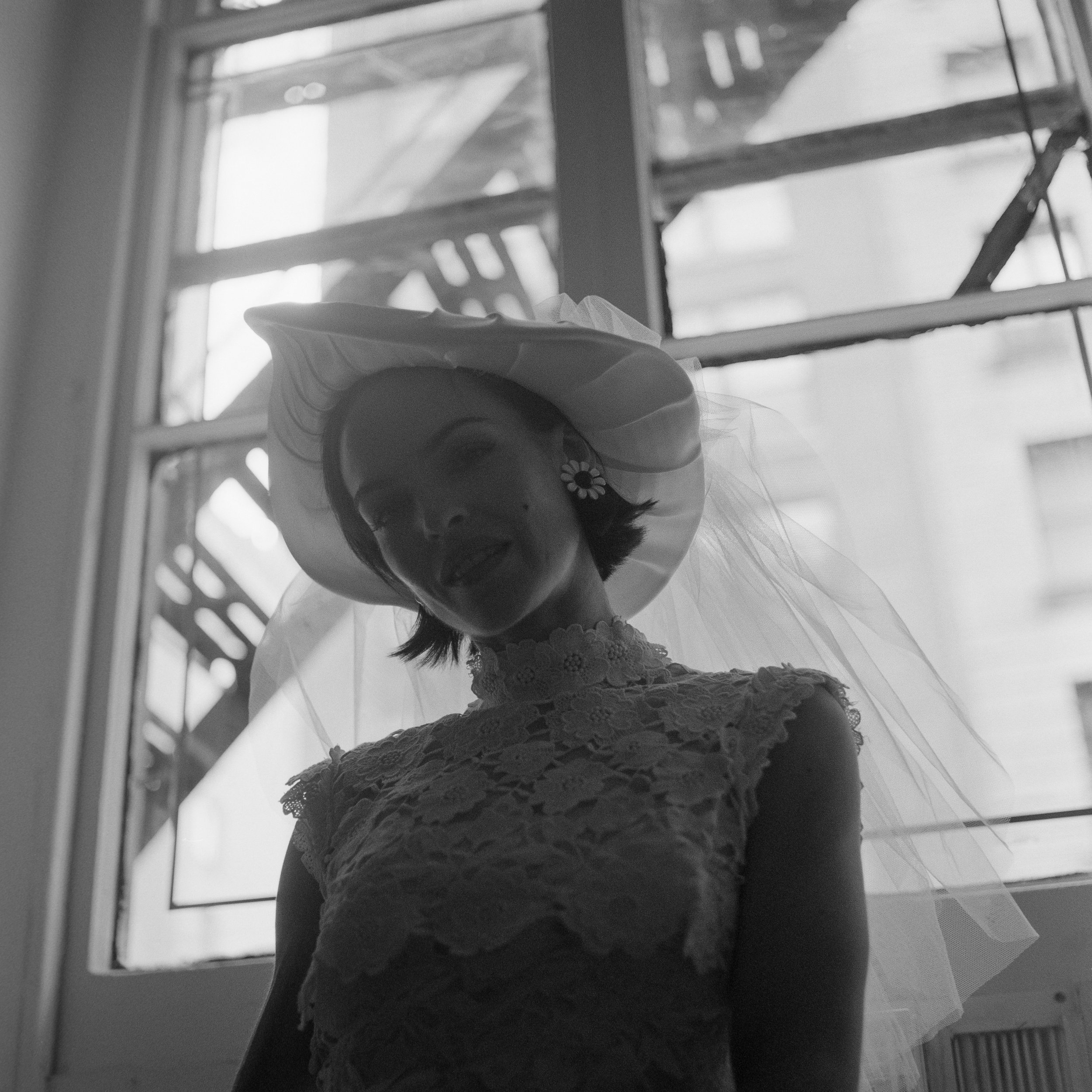 A black-and-white photo of a person standing in front of a window, wearing a lace dress with ruffles and a floral hair accessory, with the person's face partially visible and a veil over their head.