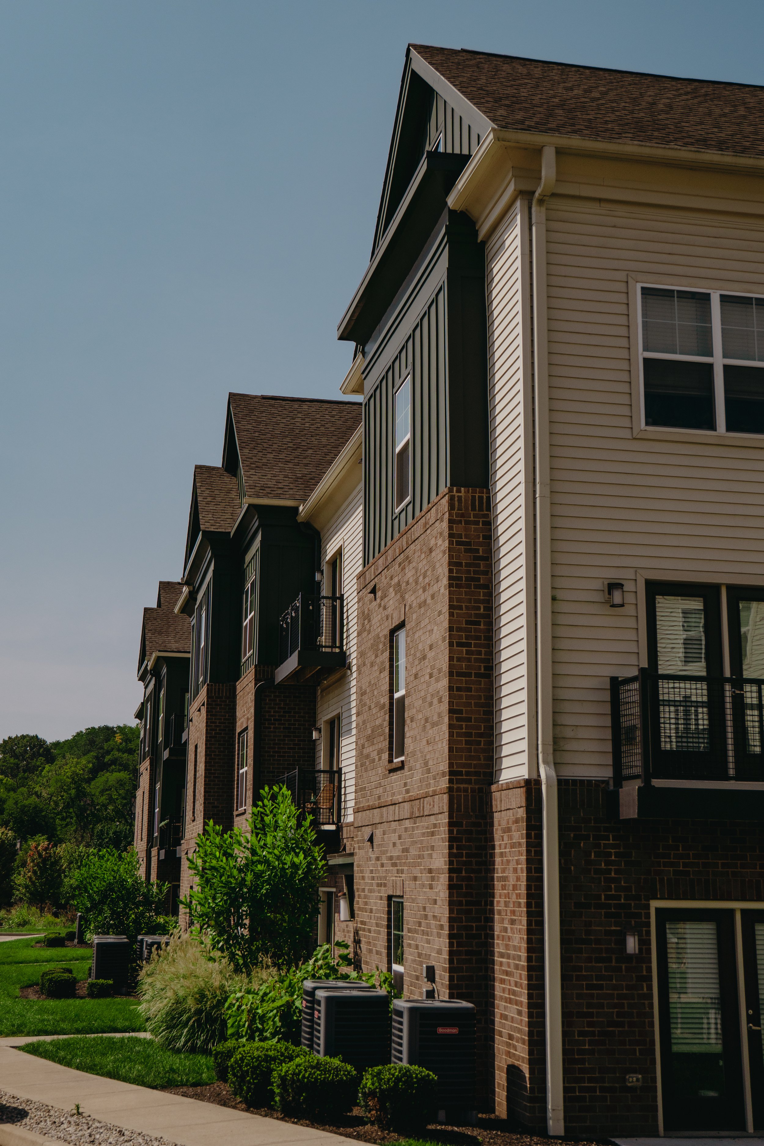 Front view of modern multi-story residential buildings with brick and siding exteriors, balconies, and well-maintained landscaping under a clear sky.