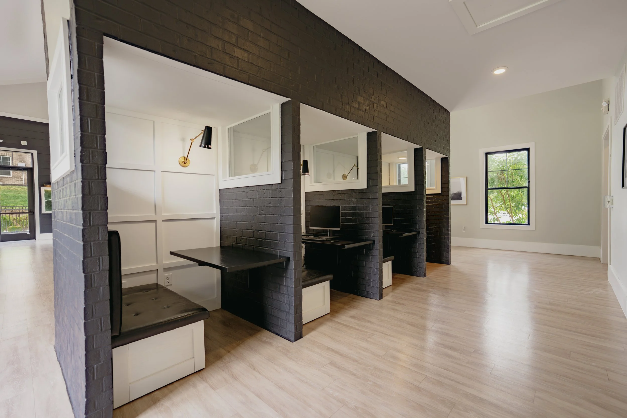 Modern interior space with black brick partitions, white walls, a computer desk, and windows showing green trees outside.