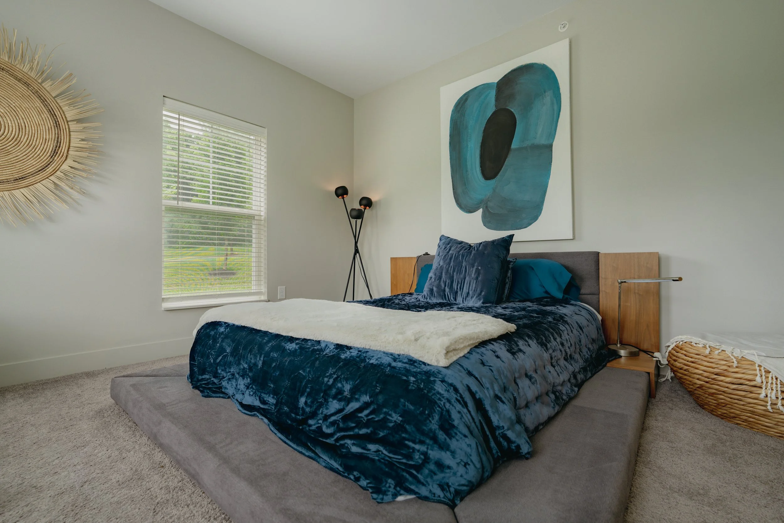 A bedroom with a bed covered in dark blue velvet bedding, a large abstract painting with blue and black shapes on the wall, a wooden nightstand, a black floor lamp, a wicker basket, a window with white blinds, and a large woven sunburst wall decor.