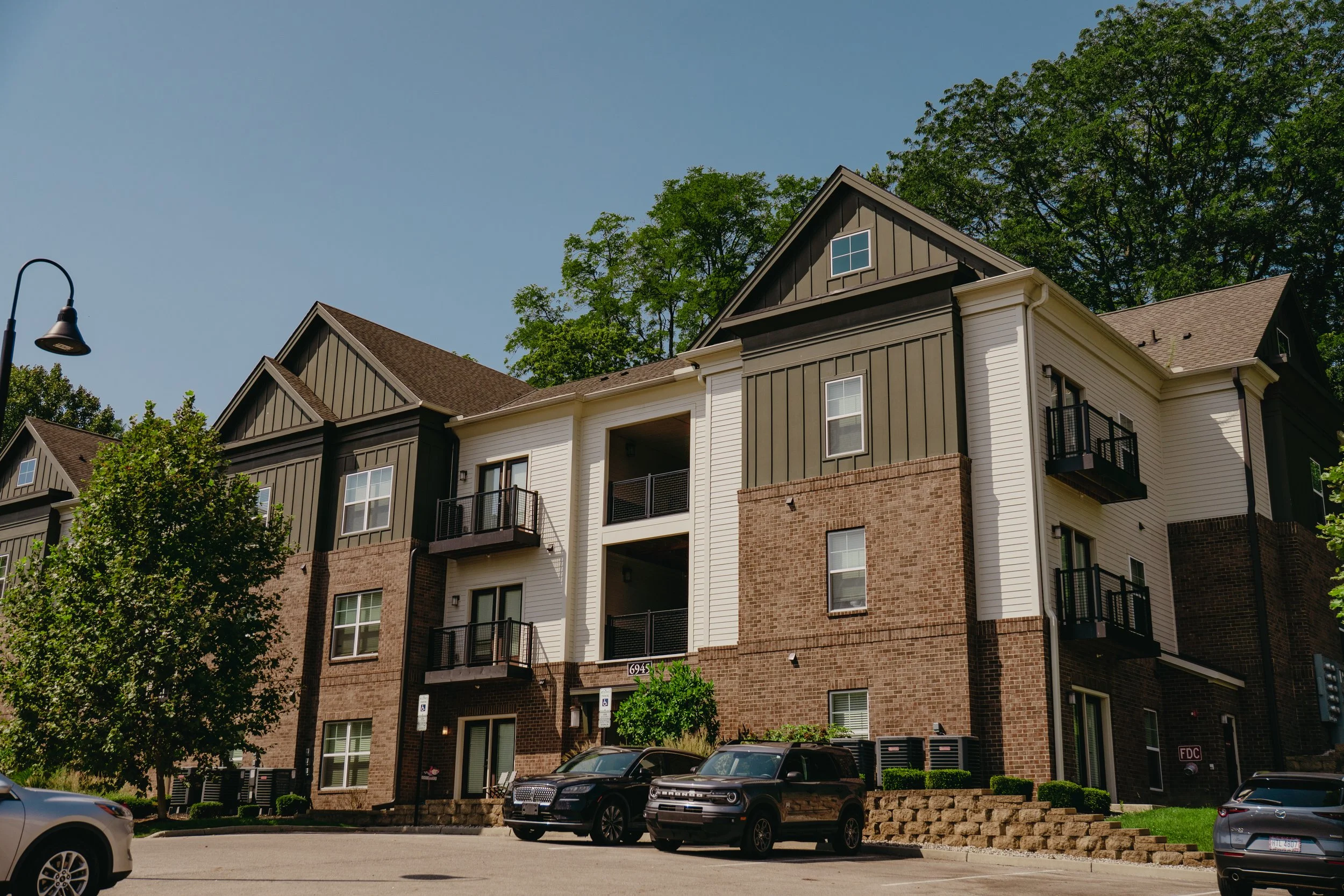 A multi-story residential building with modern architecture, featuring brick and panel siding, balconies with black railings, and several cars parked in front. There are trees and a streetlamp in the scene under a clear blue sky.