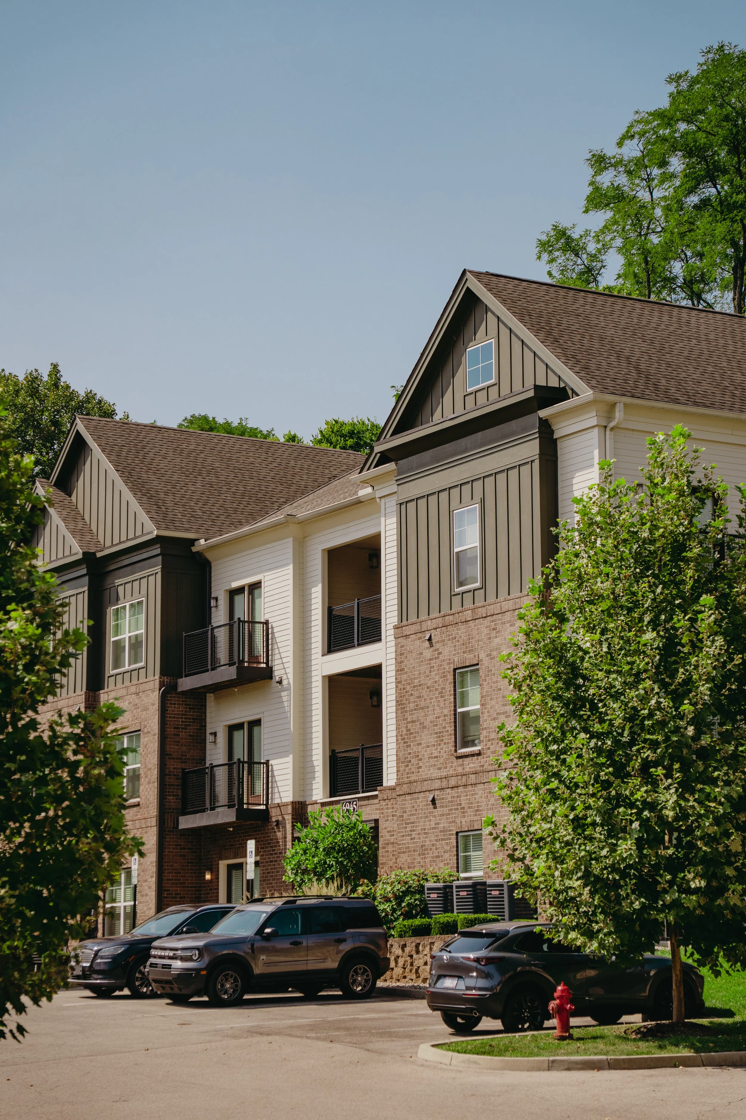 Multi-story residential building with brick and siding exterior, dormer windows, and surrounding parking lot with cars and greenery.