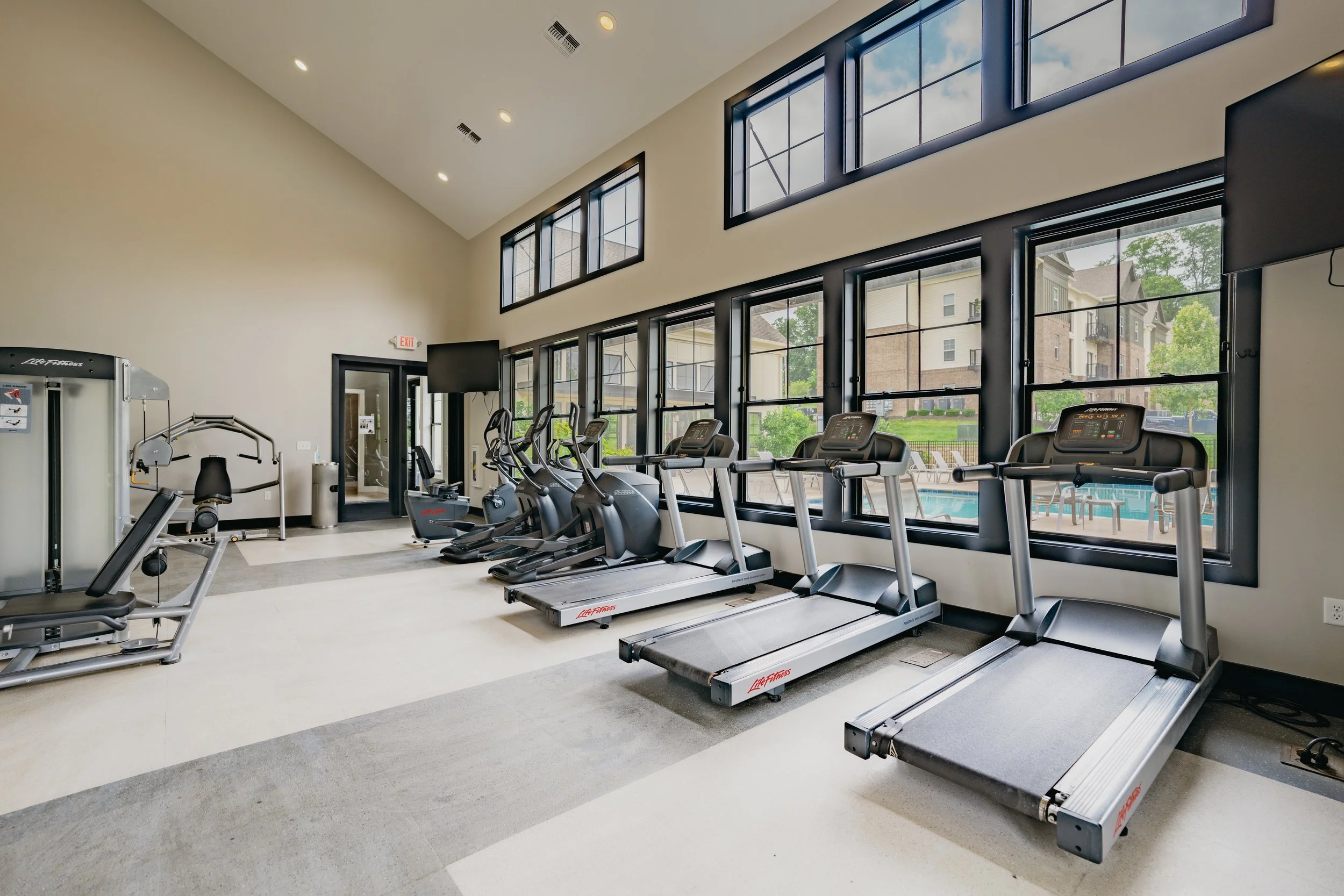 A modern gym with exercise equipment including treadmills, elliptical machines, and weight machines, large windows with a view of residential buildings and greenery, high ceilings, and neutral-colored walls.