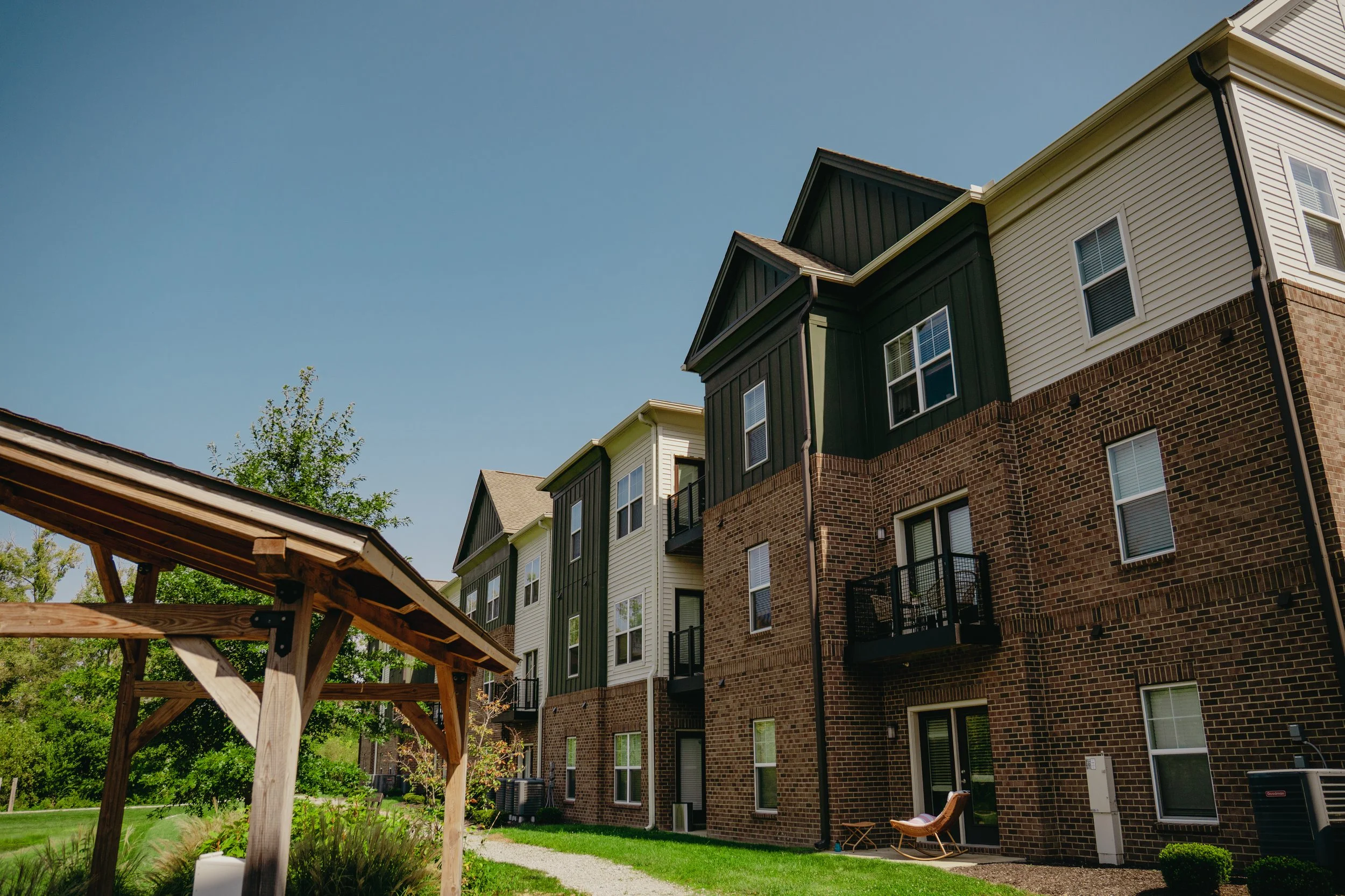 View of multi-story residential buildings with brick and siding exteriors under a clear blue sky, with a wooden pergola and green lawn in the foreground.
