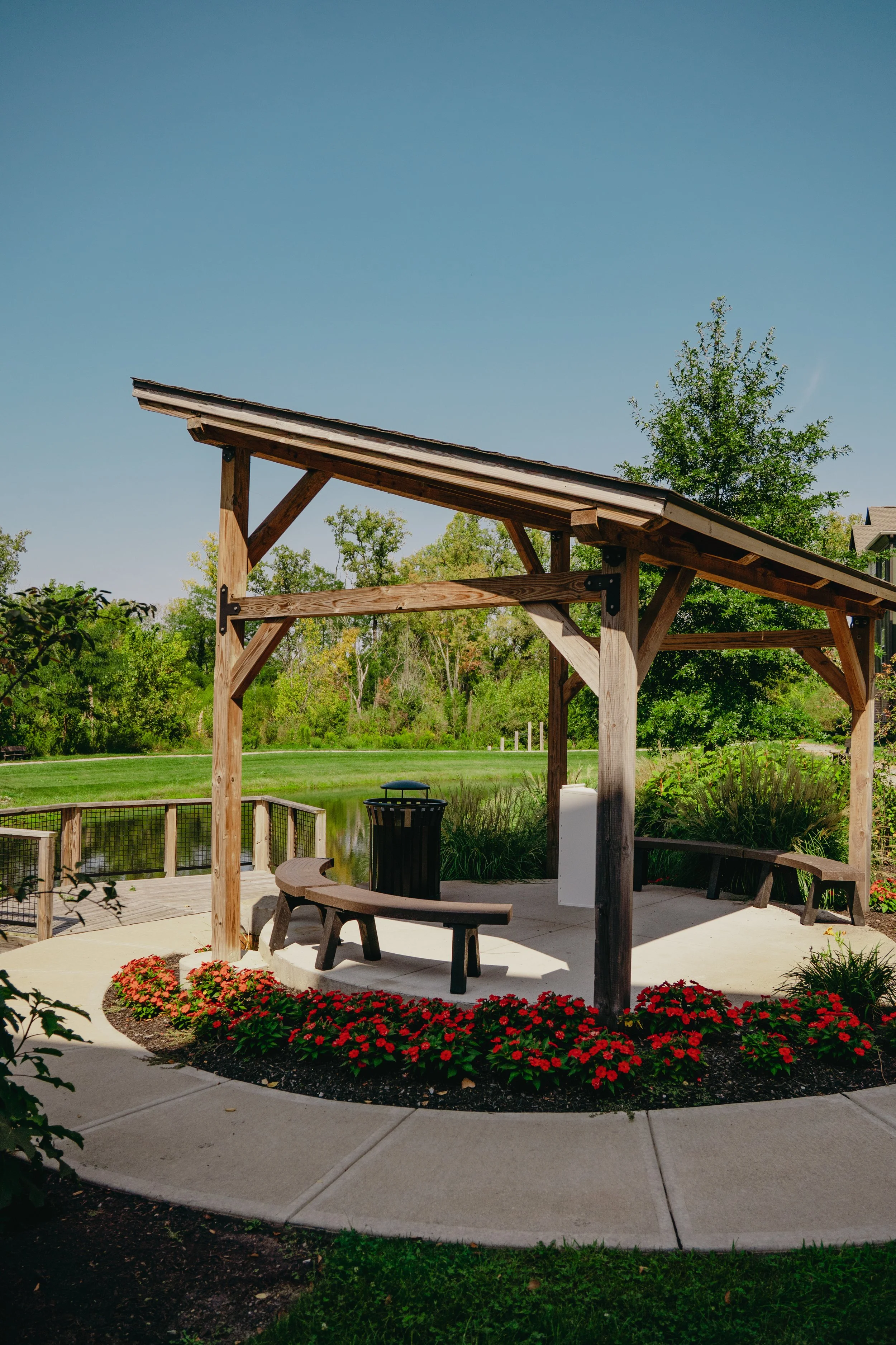 A wooden pergola with a sloped roof, situated on a patio beside a pond and a backdrop of lush green trees and a clear blue sky.