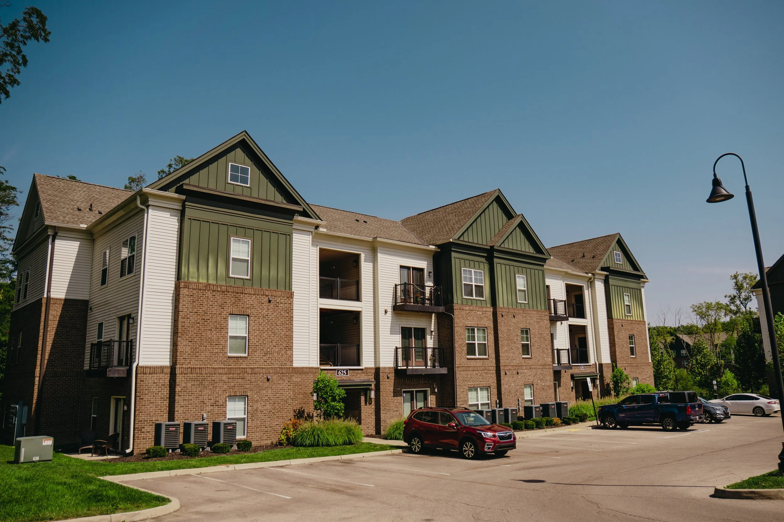 A multi-story residential apartment building with a brick and green siding exterior, several balconies, and a parking lot with parked cars in front. There is a streetlamp and some small trees and bushes near the building.