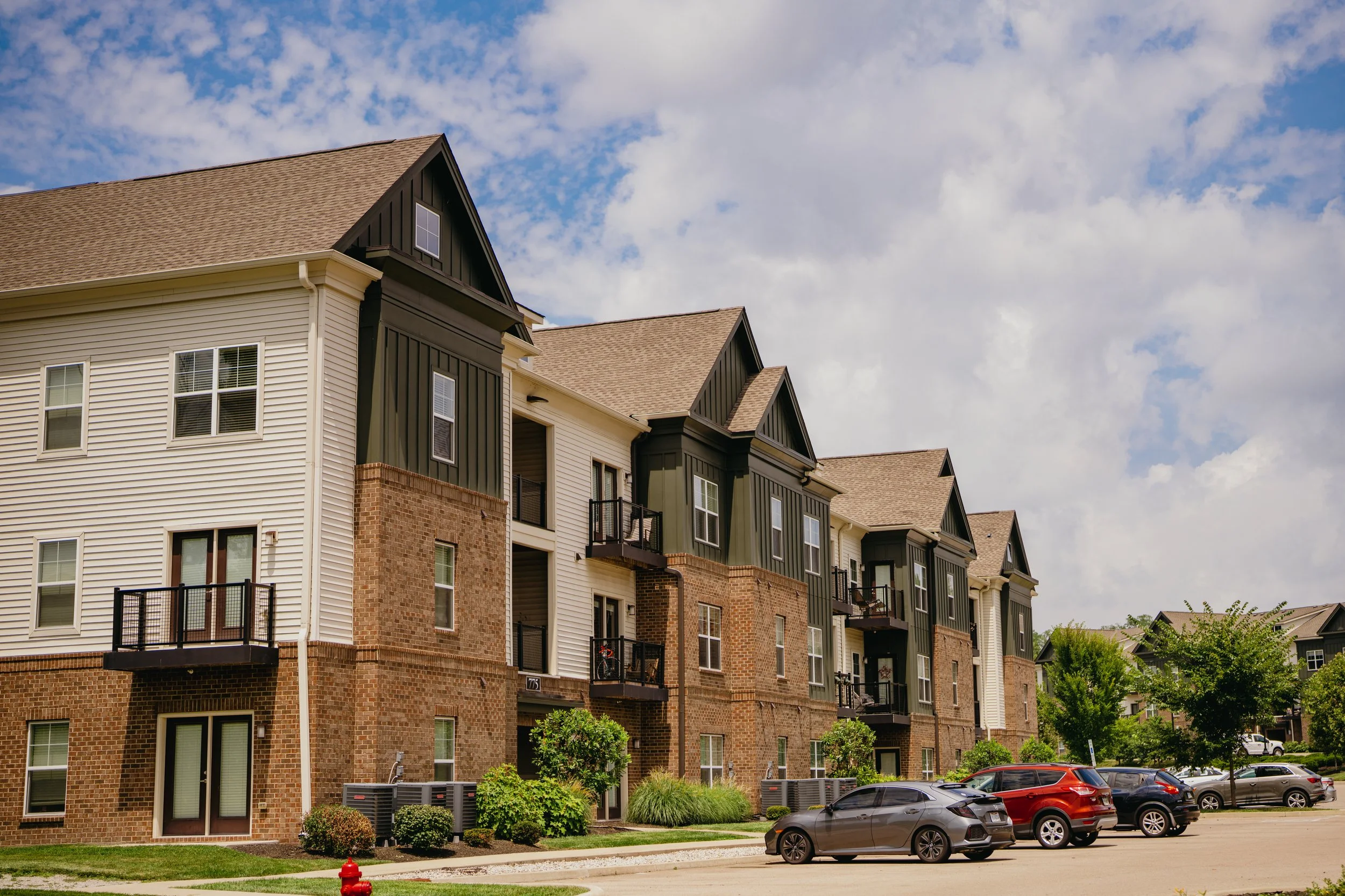 A row of multi-story residential buildings with brick and siding exteriors, multiple windows, and small balconies, under a partly cloudy sky. There is a parking lot with parked cars in front, green landscaping, and a fire hydrant on the sidewalk.