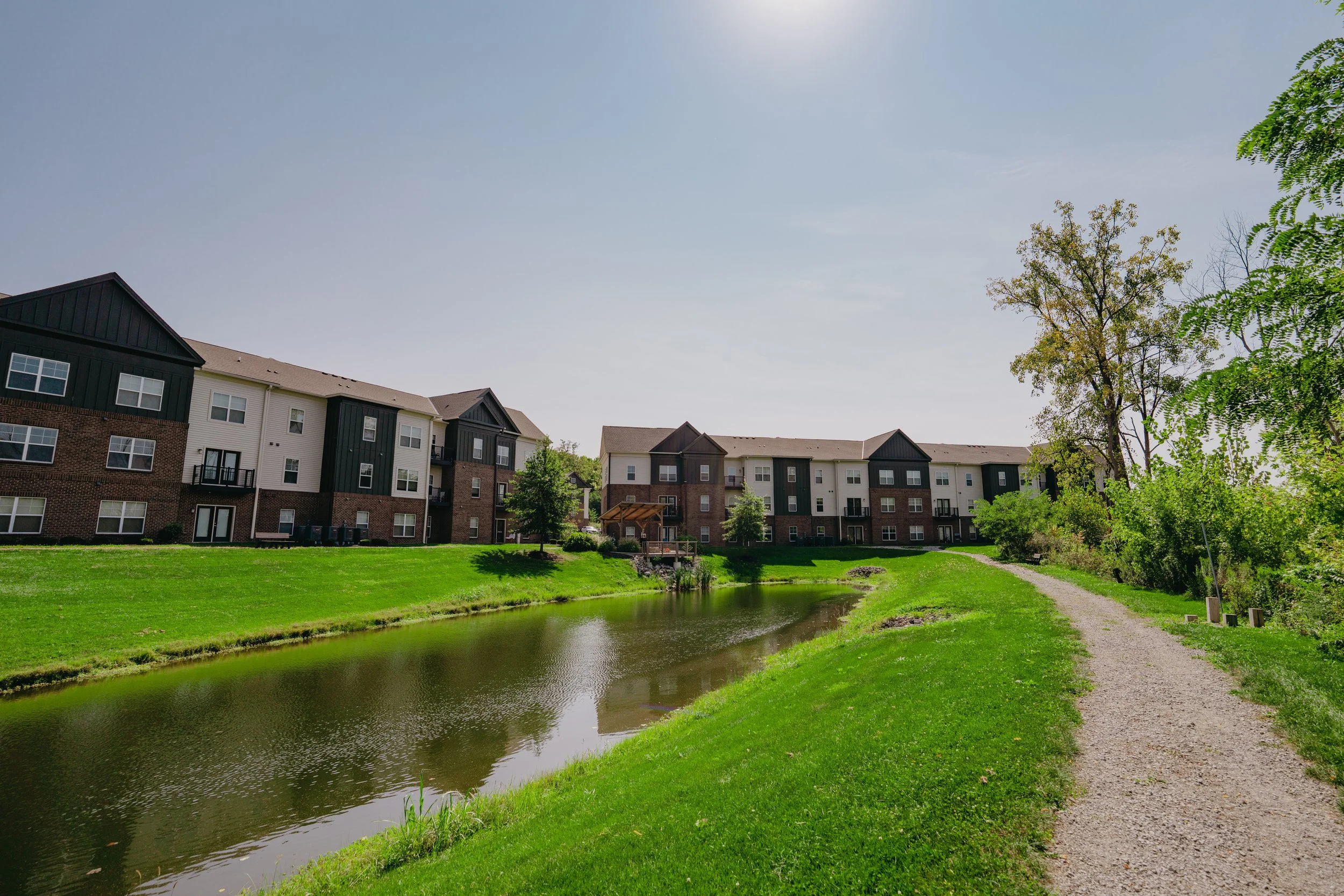 A peaceful residential area with apartment buildings, a small pond, a gravel walking path, and green trees under a clear blue sky.