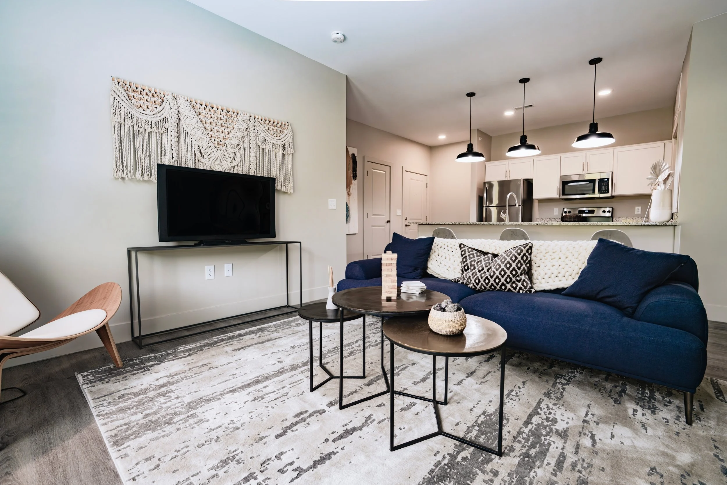 Modern living room with navy blue sofa, patterned pillows, two round coffee tables, and a TV on a black metal stand. Wall decor is a white macrame hanging. The space features a high ceiling with three black pendant lights and open kitchen with white cabinets, granite countertops, and stainless steel appliances.