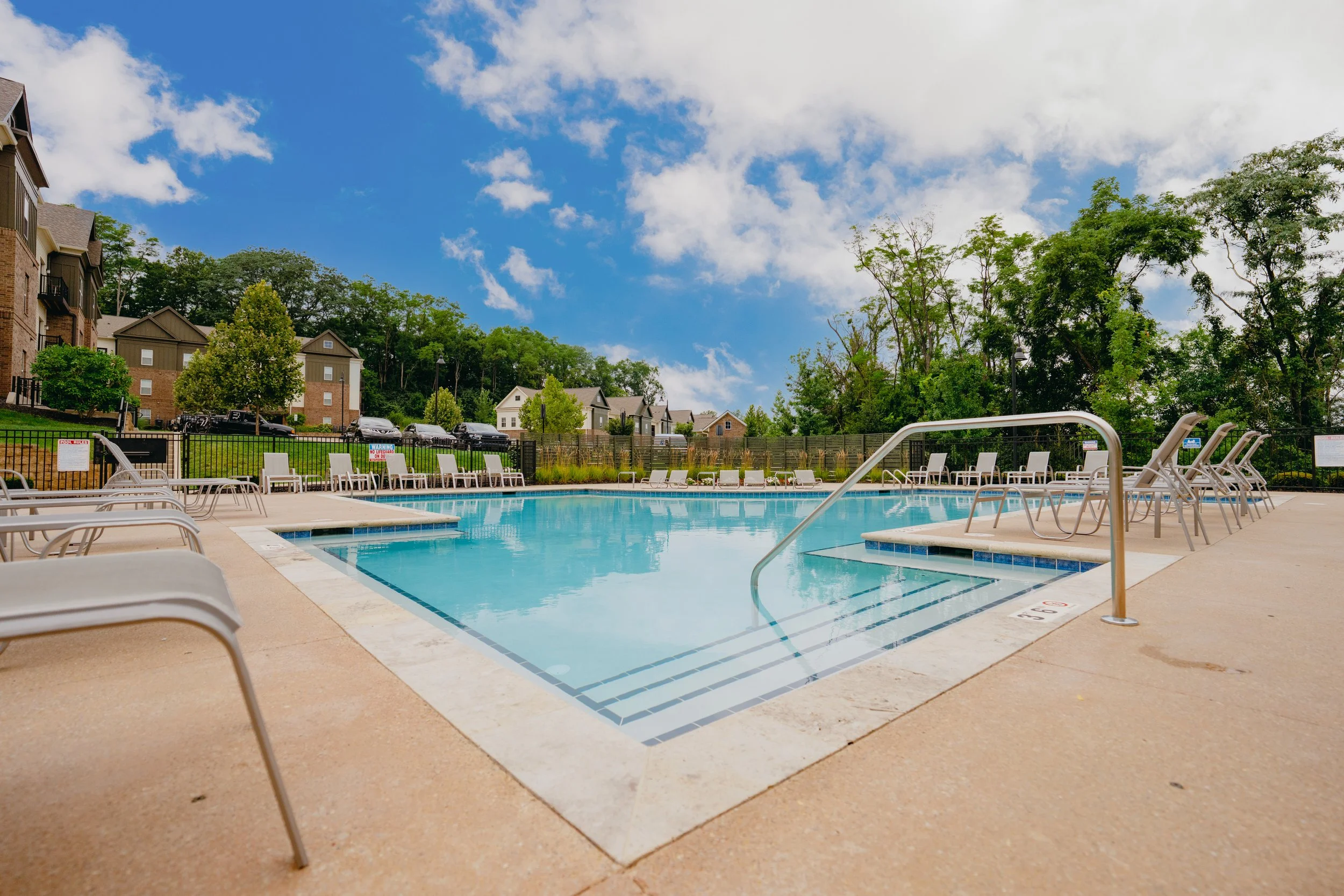An outdoor swimming pool surrounded by lounge chairs on a concrete deck, with a black metal fence and residential buildings in the background, under a partly cloudy blue sky with green trees.