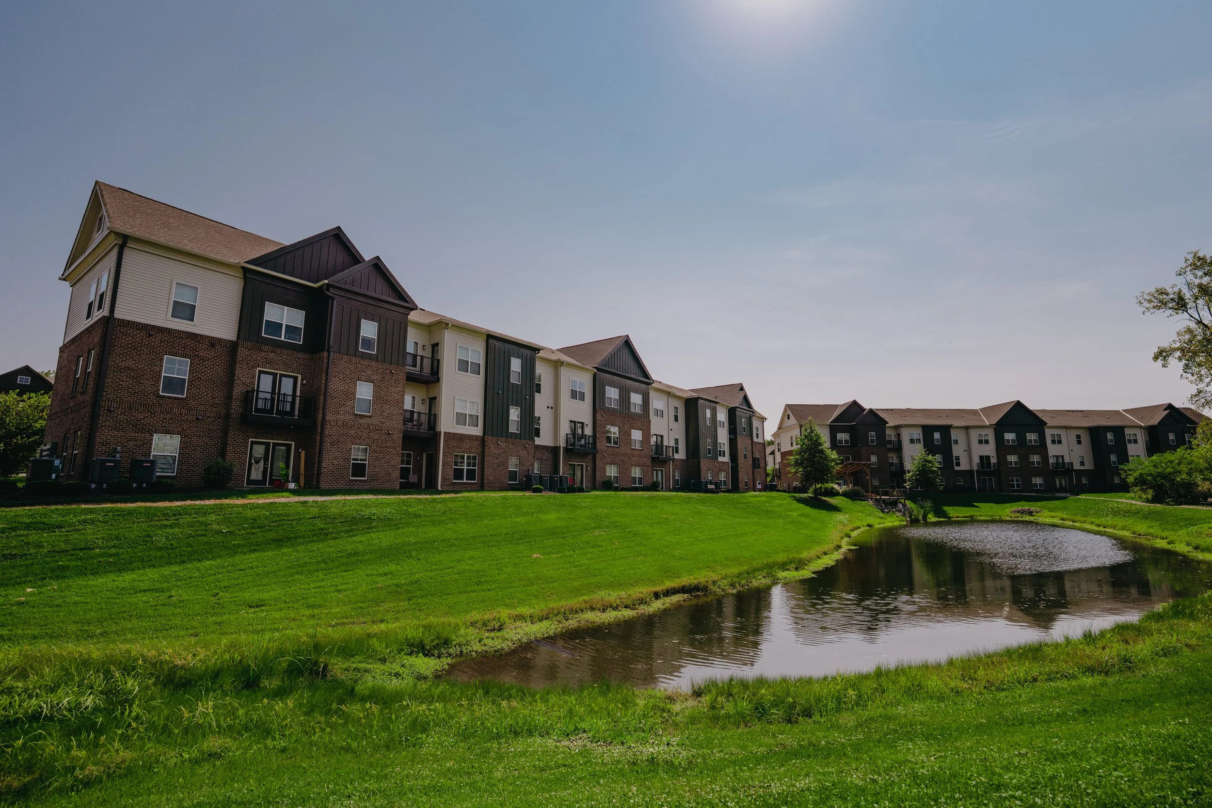 A multi-story residential apartment complex with brick and siding exterior, surrounded by green grass and small trees, next to a pond under a clear blue sky.