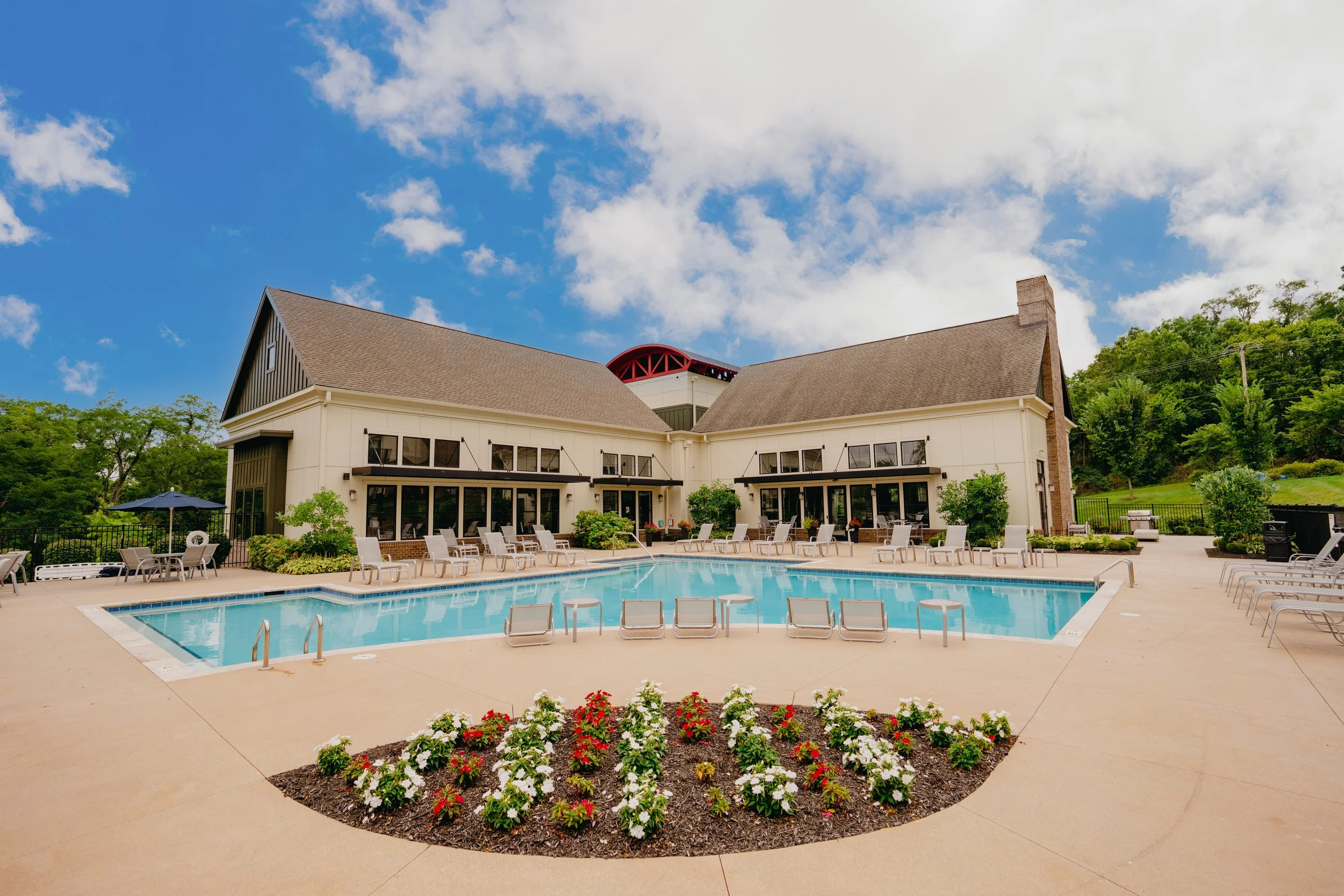 A backyard swimming pool surrounded by lounge chairs with a building in the background, clear blue sky with clouds, and a flower bed in the foreground.