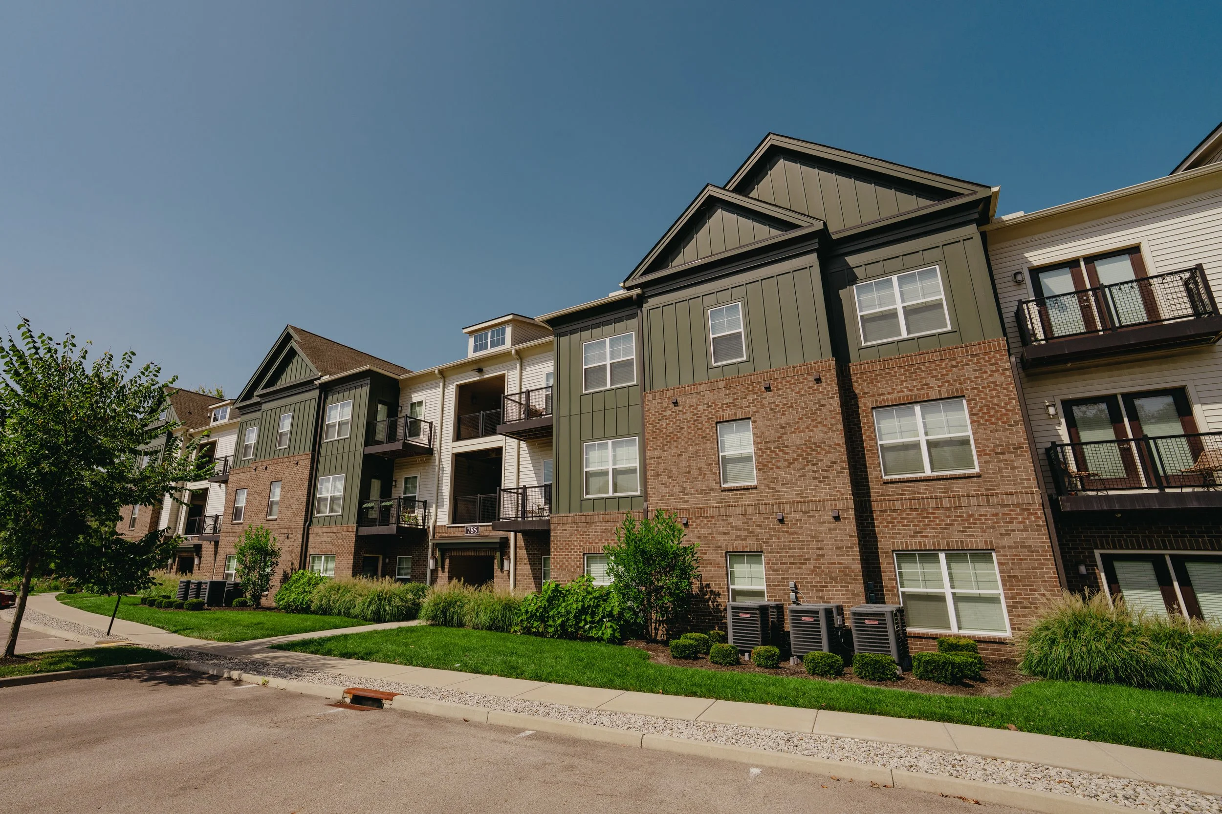 A modern multi-story apartment complex with brick and green siding, featuring multiple windows and small balconies, surrounded by green grass, trees, and landscaped plants under a clear blue sky.