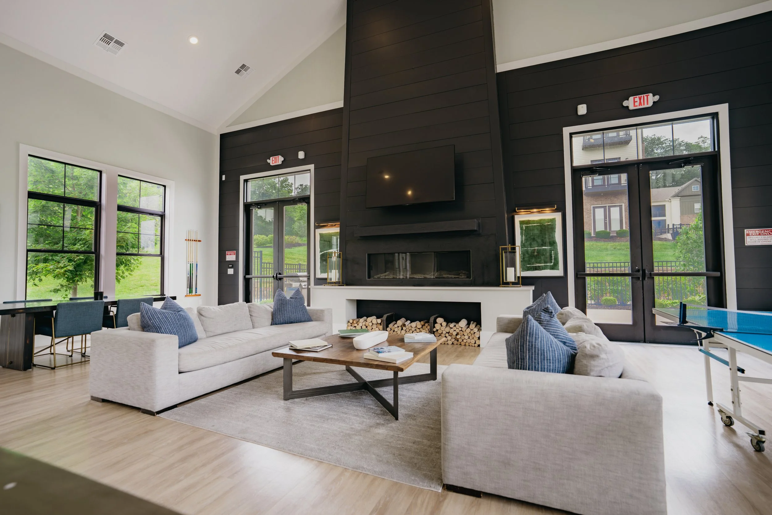 A spacious room with large windows, a white sofa with blue pillows, a wooden coffee table with books, and a modern fireplace with a mounted TV above it. There is a black wall and greenery visible outside.