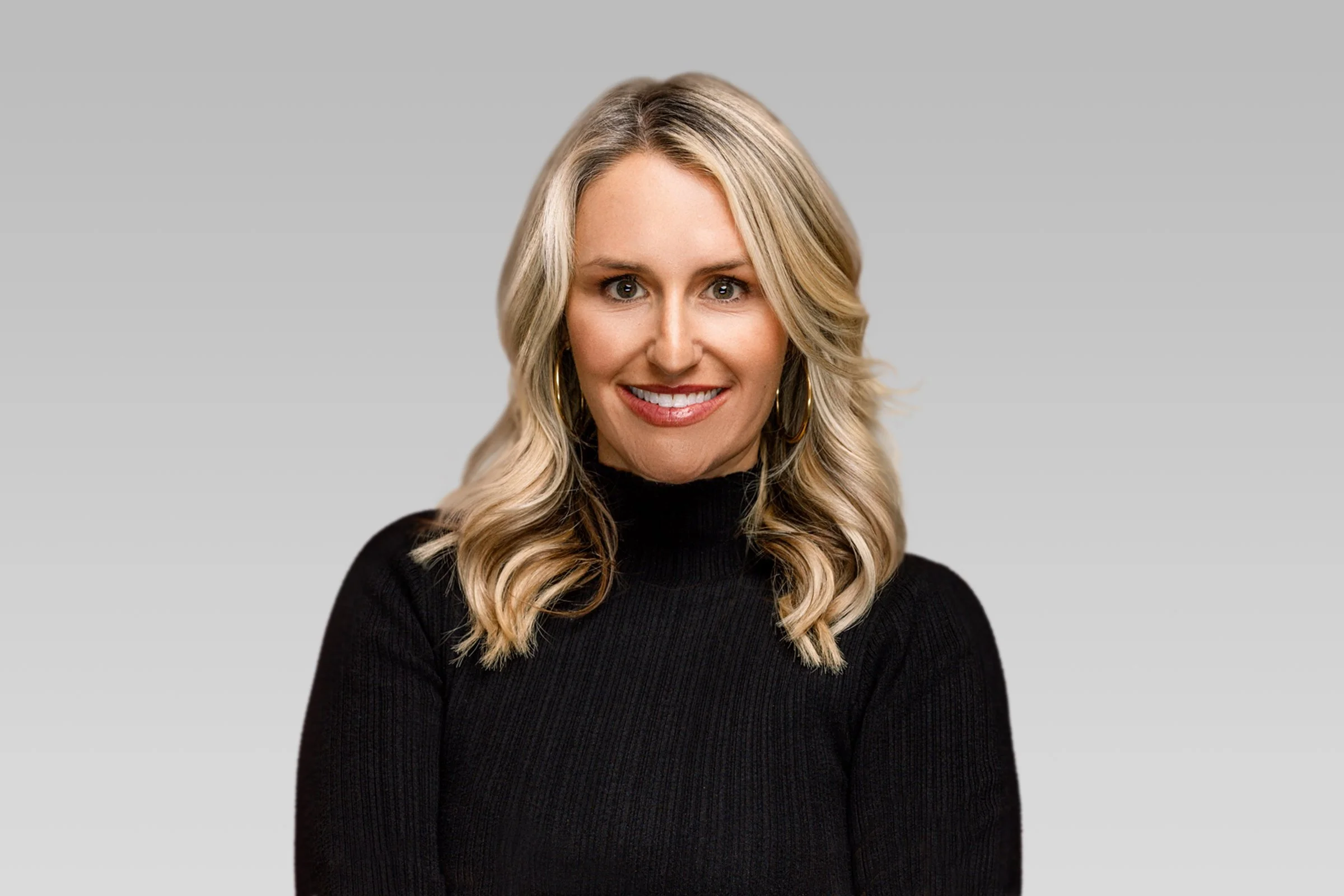 Portrait of a woman with blonde hair in loose waves, wearing a black turtleneck top and large hoop earrings, smiling against a plain gray background.