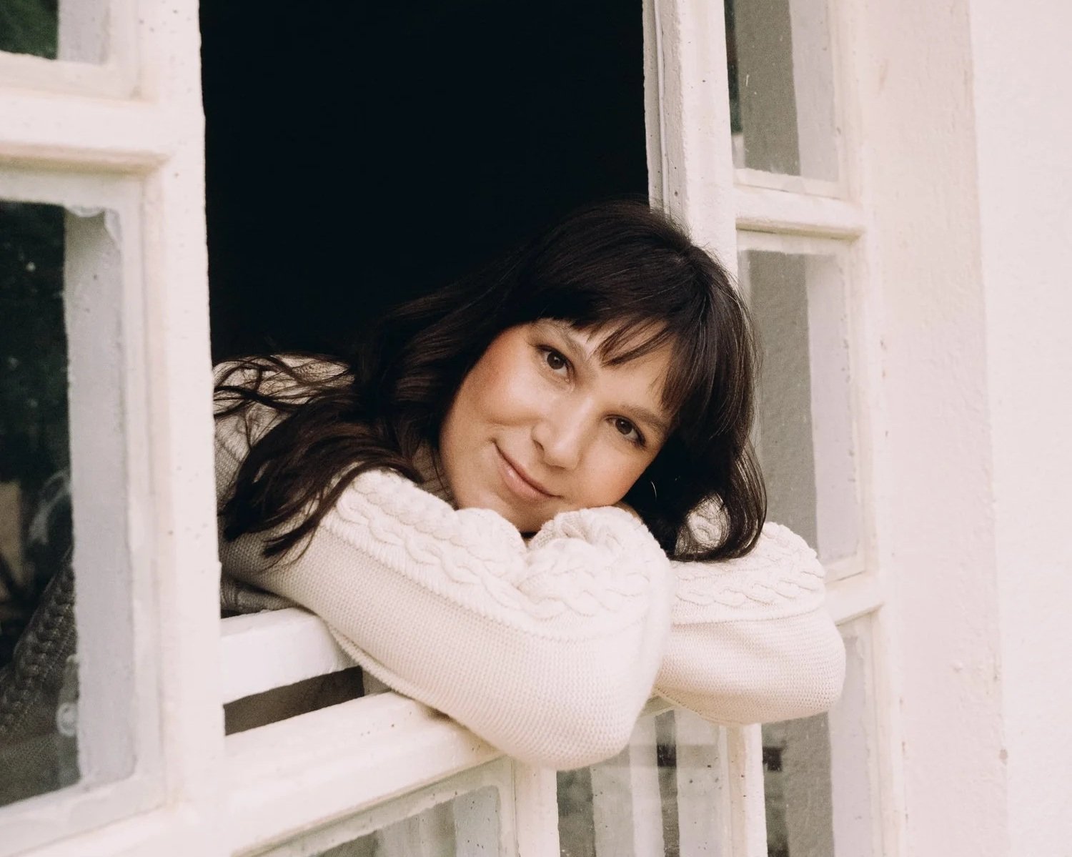 Woman with dark hair leaning on a windowsill, smiling, wearing a cream-colored sweater, with white wall and window panes in the background.