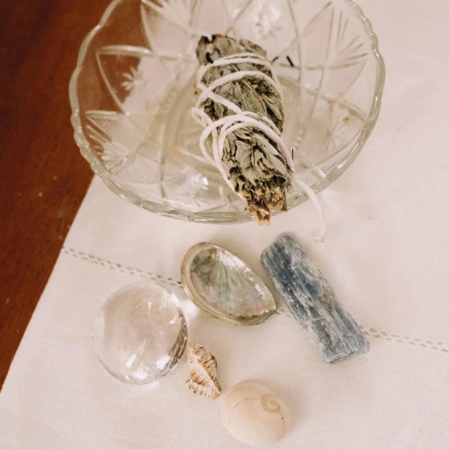 A glass bowl containing a bundle of dried herbs tied with string, placed on a white lace tablecloth. Surrounding the bowl are various seashells, a clear glass sphere, and a small piece of coral. A rustic candle holder with a blue-gray patina is also present.