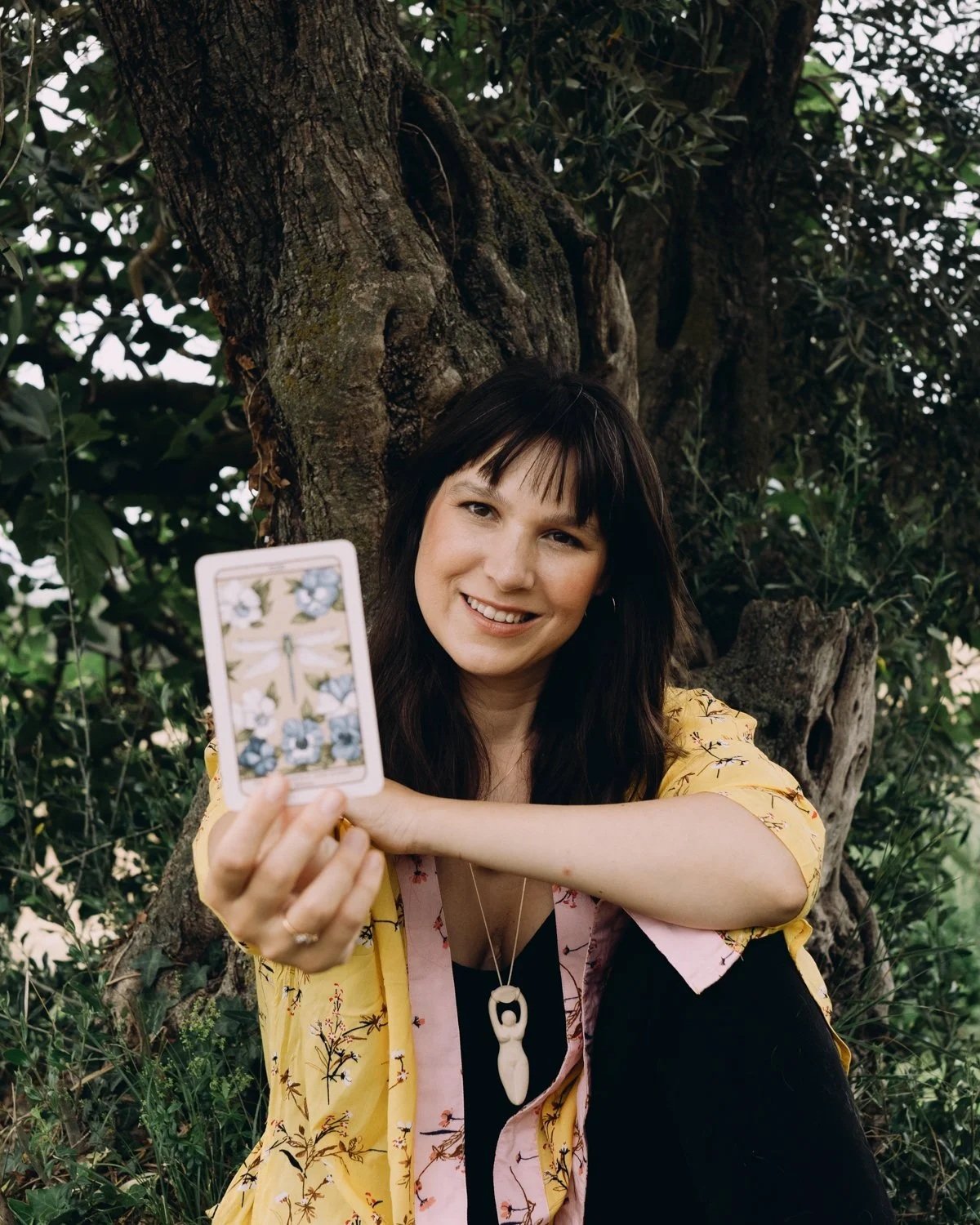 A woman sitting outdoors near a tree, holding a tarot card with floral artwork towards the camera and smiling.
