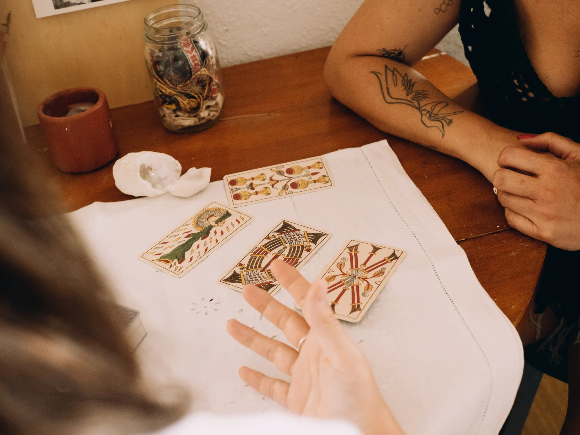 A tarot card reading session with three visible tarot cards spread on a white cloth on a wooden table. One person's hand is gesturing towards the cards, and a second person is sitting across with a tattooed arm visible. A jar filled with various cords and a small pot are on the table along with a crumpled tissue and a rolled tissue.
