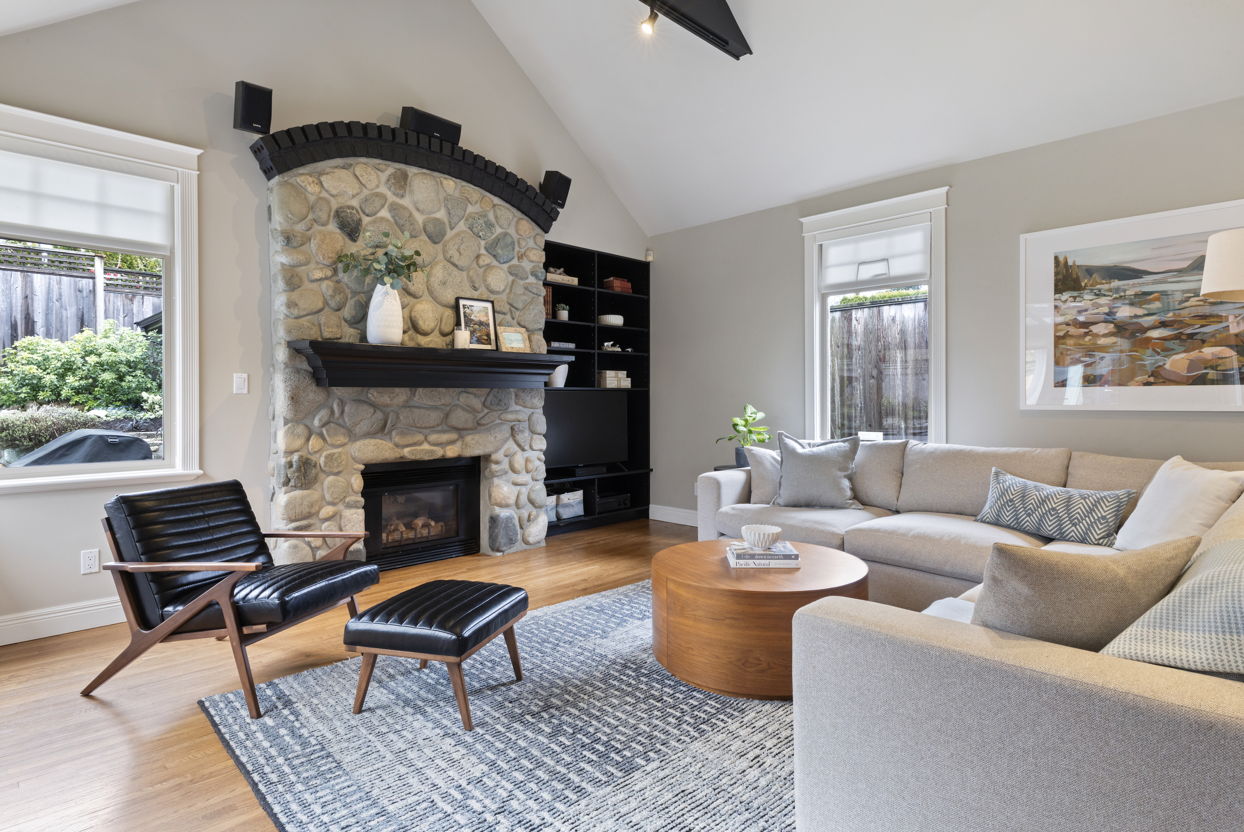 Living room with a stone fireplace, black speakers on the mantel, a black bookshelf, a beige couch with pillows, a round wooden coffee table, a black leather chair with wood armrests, and large windows with white trim letting in natural light.