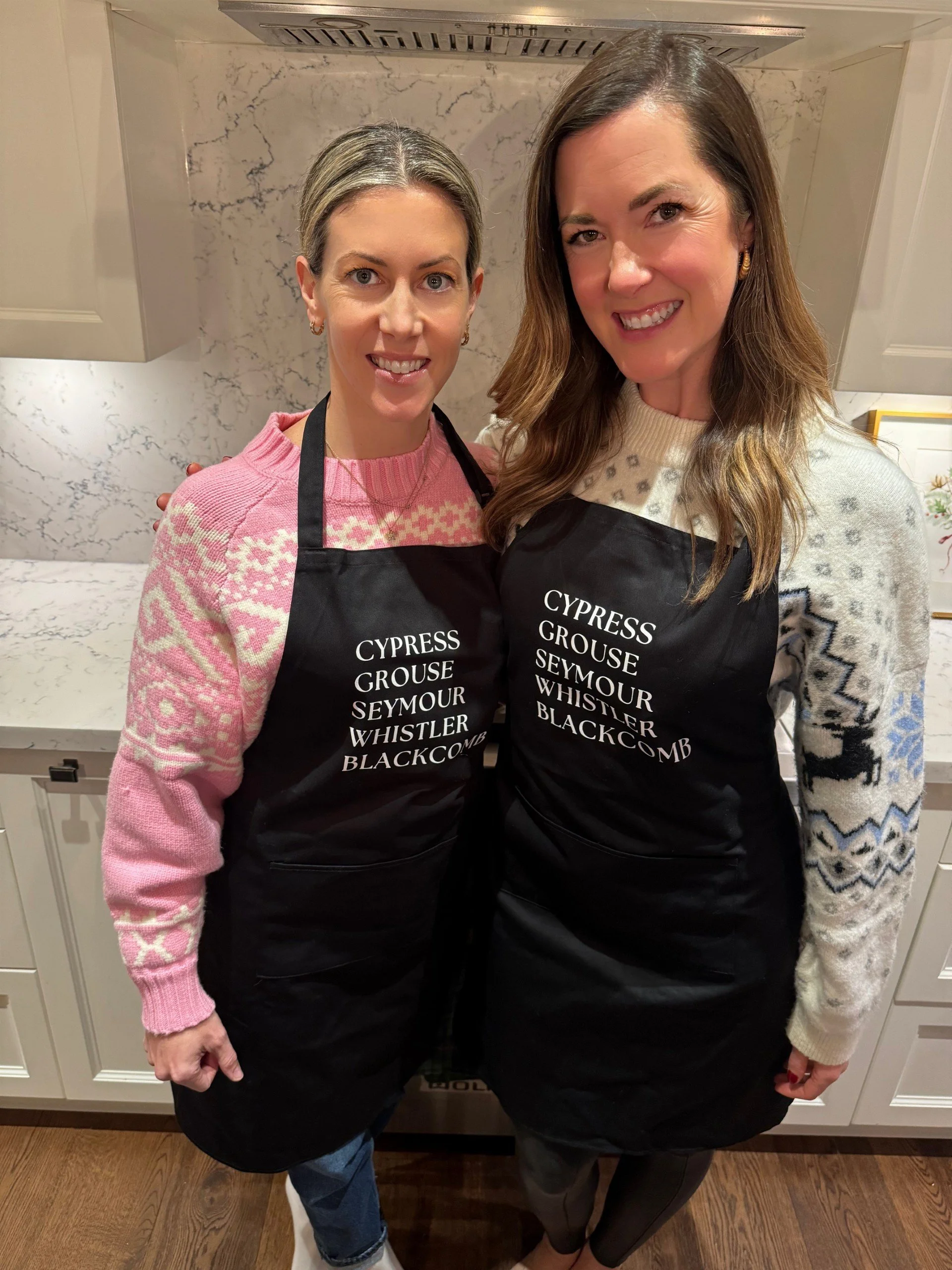 Two women standing in a kitchen, wearing black aprons with white text listing different bird names, smiling at the camera.