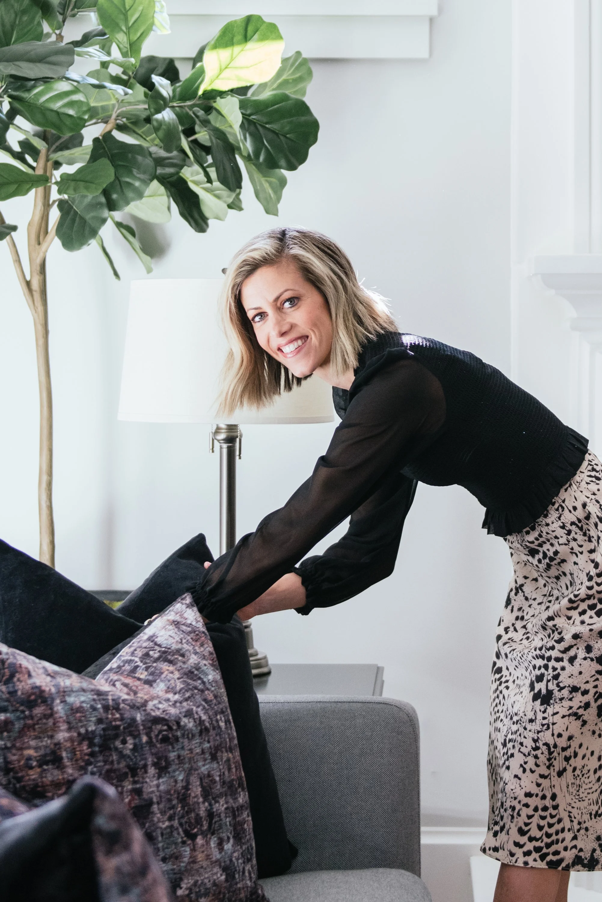 A woman with shoulder-length blonde hair smiling at the camera while bending down in a modern, well-lit room. She is wearing a black long-sleeve top and patterned skirt, with a large green potted plant and a white table lamp in the background.