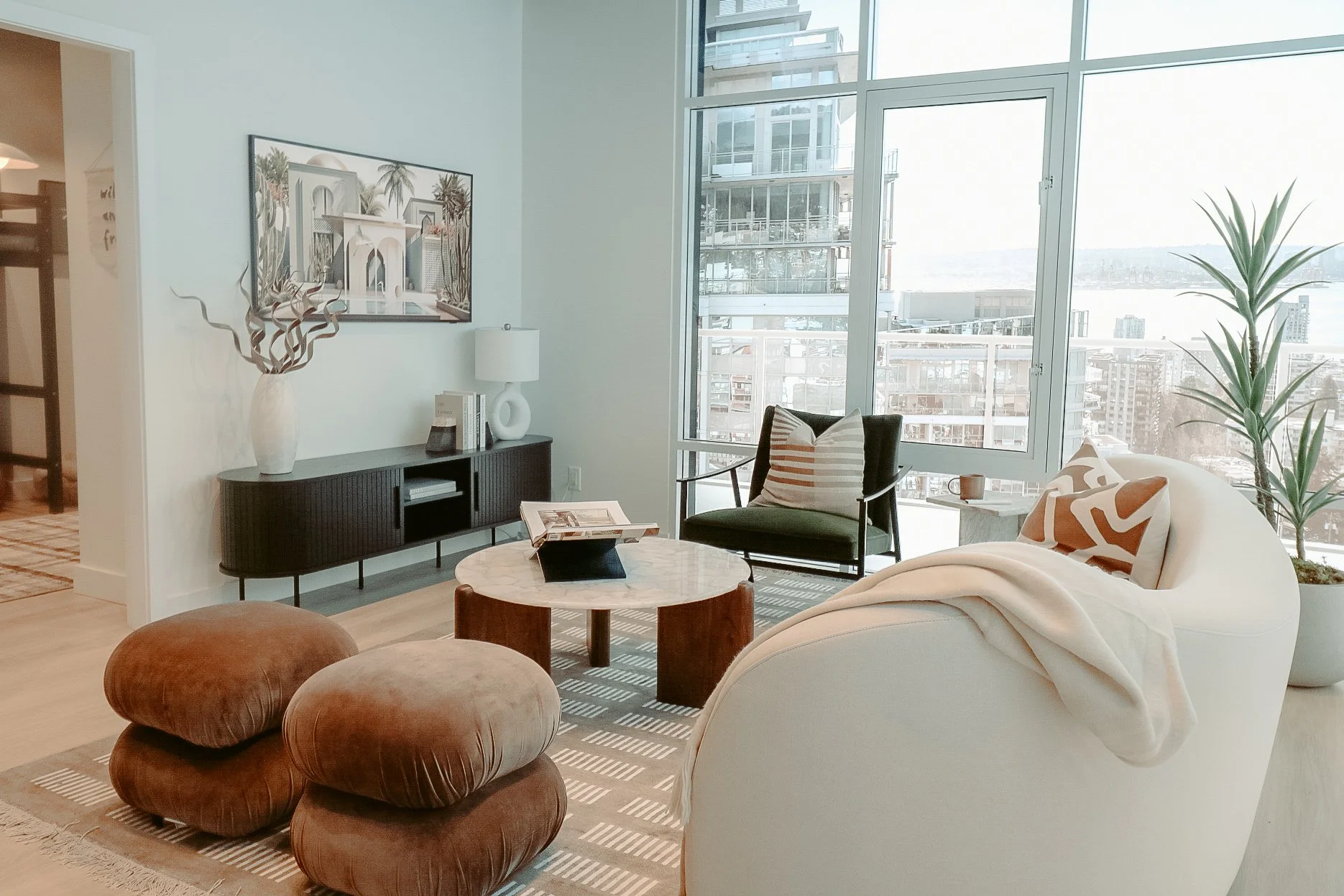 Modern living room with large windows, a black entertainment console, a white sofa with patterned pillows, a black armchair, and three brown ottomans on a striped rug, with cityscape view outside.
