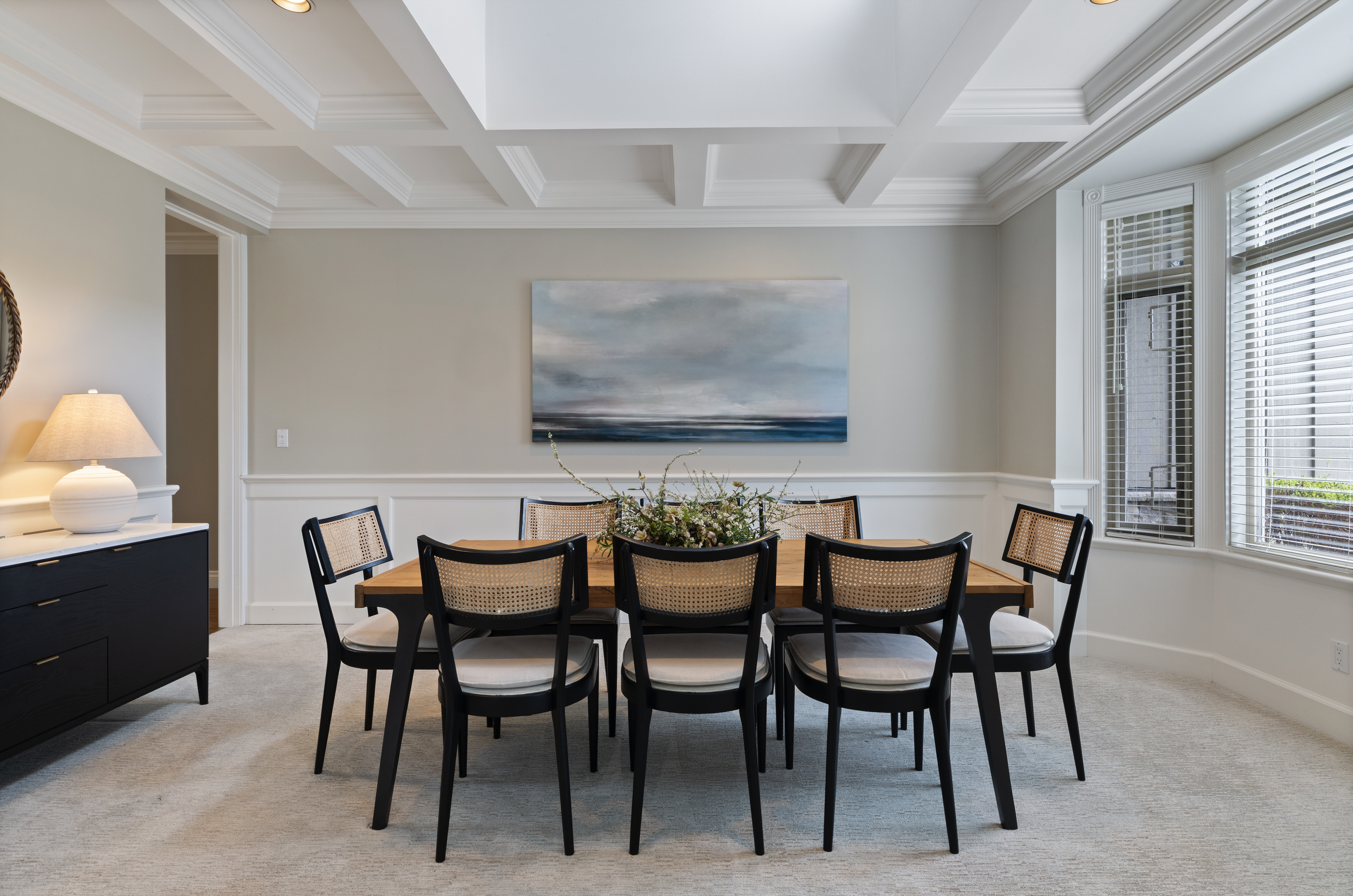 A dining room with a wooden table surrounded by eight black chairs with woven rattan backs. The room has large windows with white plantation shutters, a white wainscoting wall with a large seascape painting, a cream-colored lamp on a black sideboard, and a decorative floral centerpiece on the table.